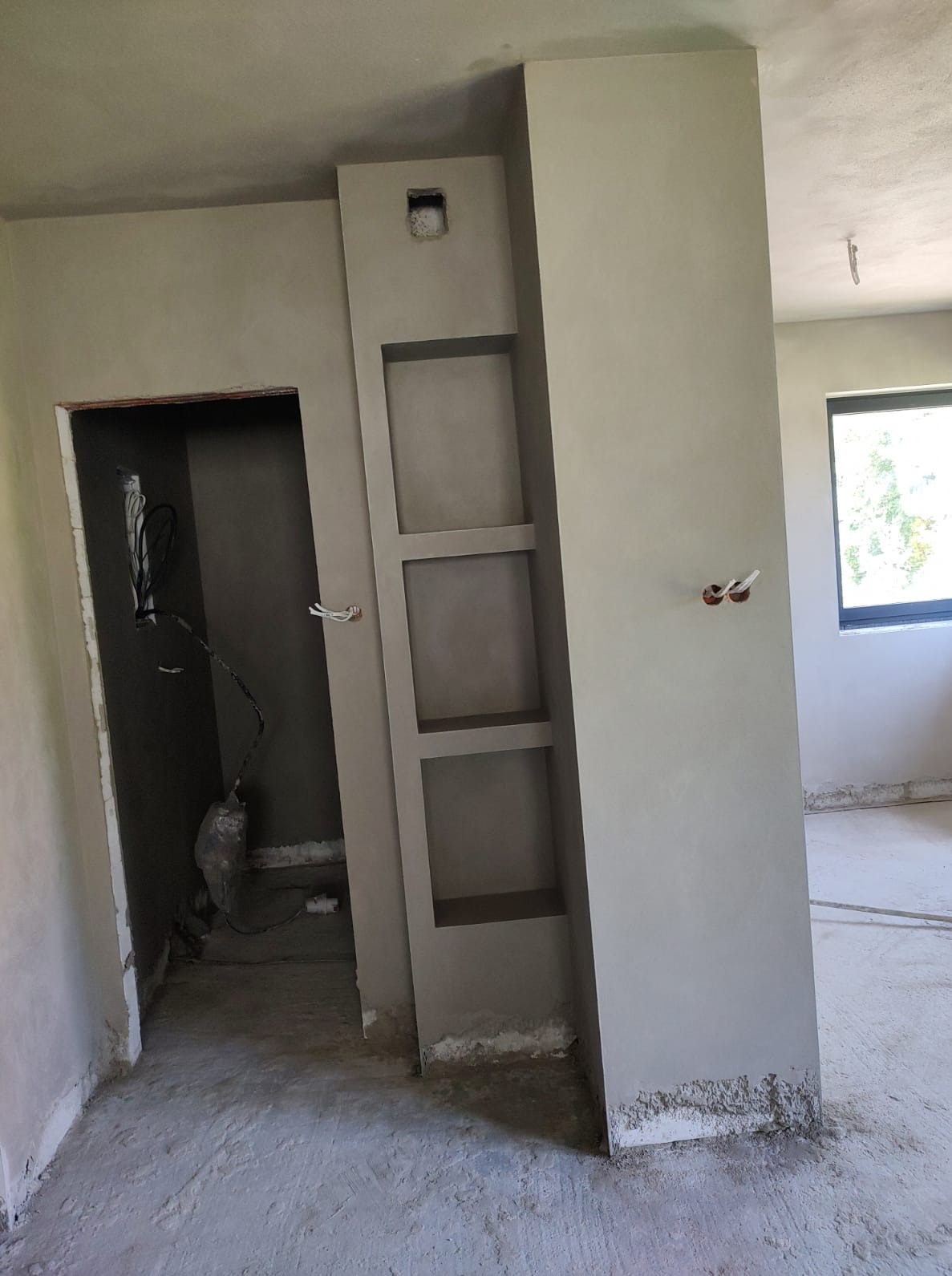 Wnętrze po tynkowaniu maszynowym: gładkie ściany w odcieniach szarości, wnęka z półkami i widoczne kable w otworze. Surowy stan wykończenia podłogi.