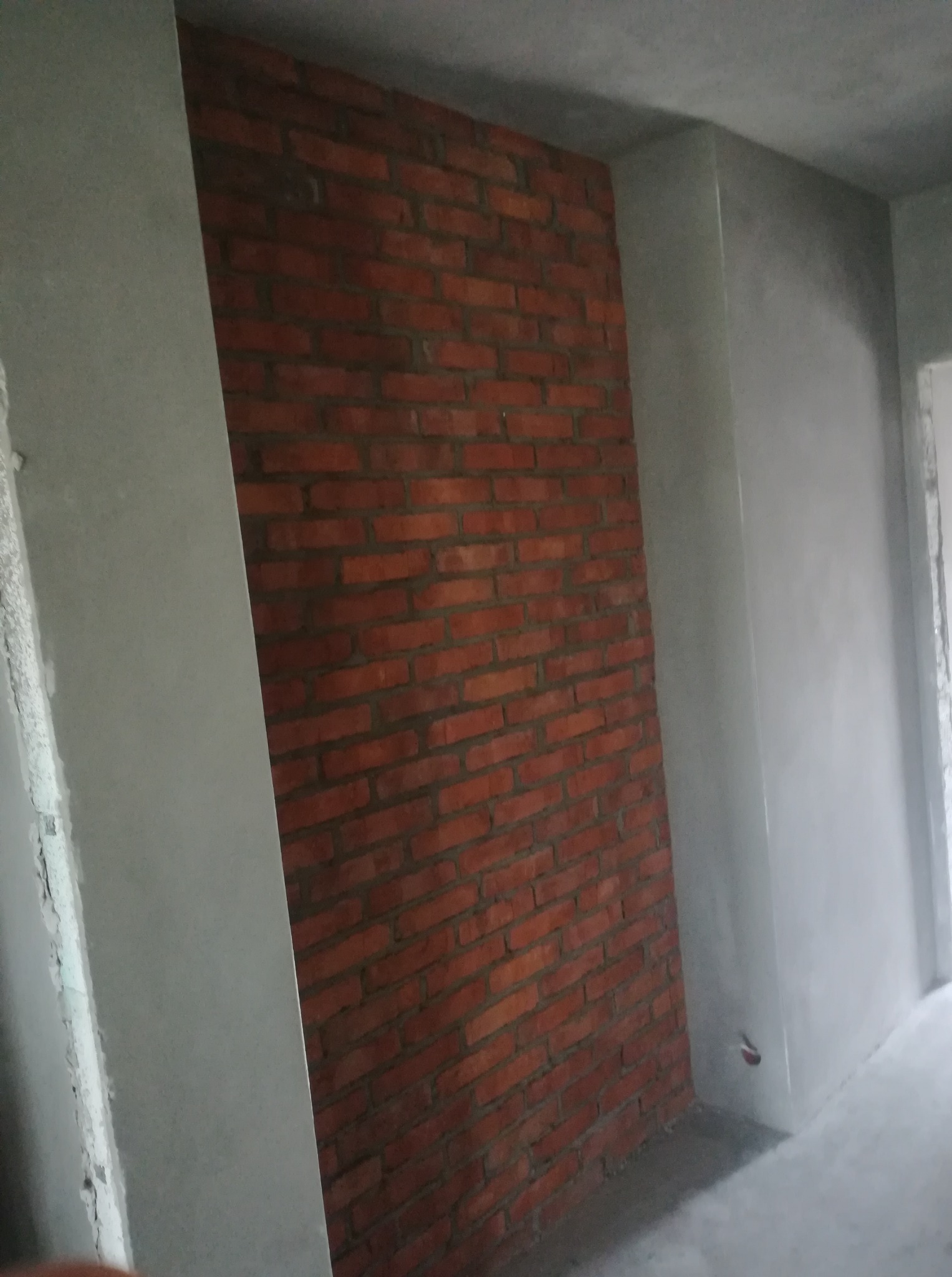 Wnętrze w trakcie prac wykończeniowych: fragment ściany z czerwoną cegłą sąsiaduje z gładką, szarą ścianą pokrytą tynkiem. Widoczny fragment podłogi i narożnik pomieszczenia.