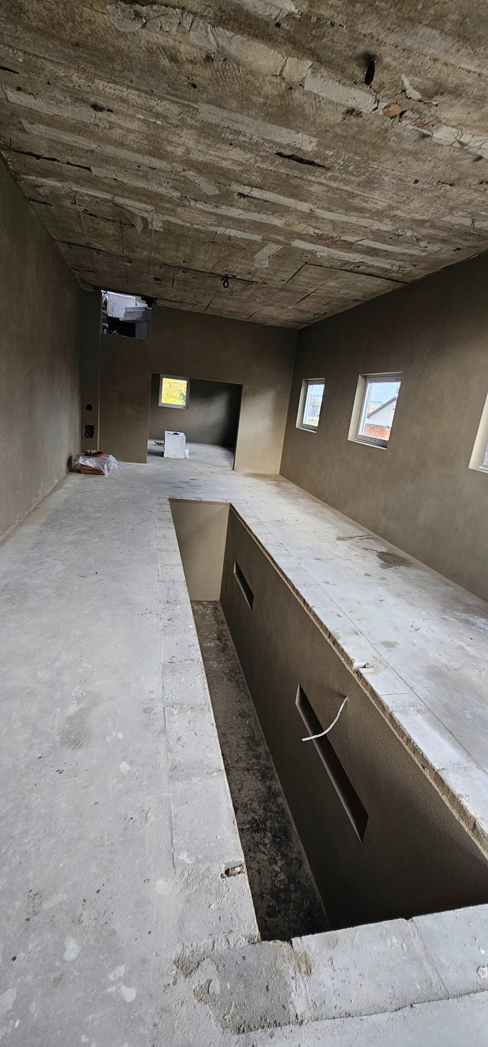 Wnętrze w trakcie prac wykończeniowych: ściany pokryte świeżym tynkiem w odcieniach szarości, surowy betonowy strop, okna wpuszczające naturalne światło. Widoczne elementy instalacji.