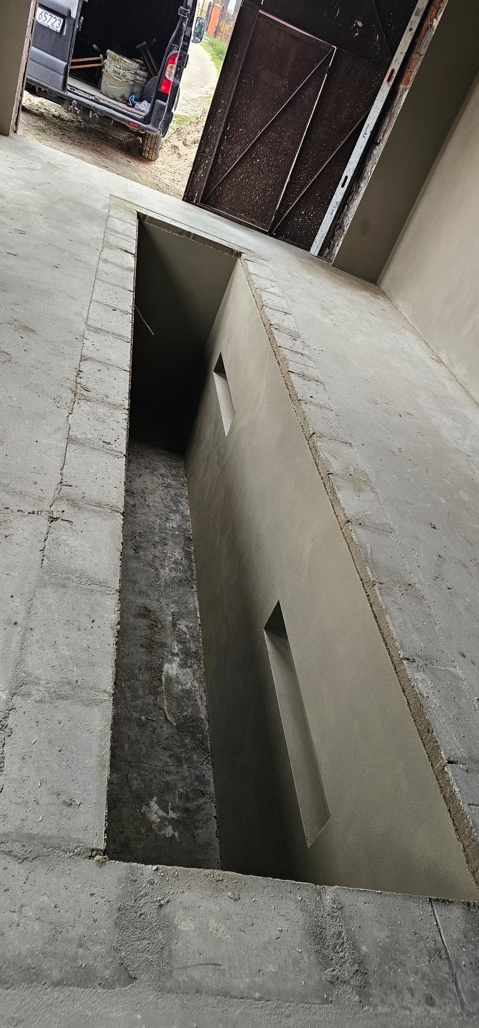 Wnętrze garażu z przygotowanym kanałem, świeżo po tynkowaniu ścian. Widoczny samochód dostawczy z otwartymi drzwiami i narzędziami w tle.