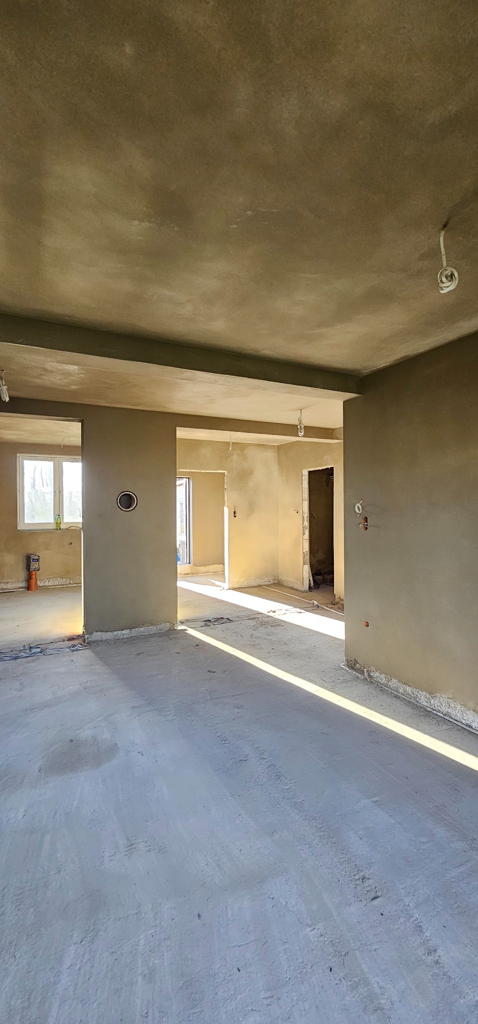 Wnętrze w stanie deweloperskim po nałożeniu tynków: szare ściany, sufit i podłoga, widoczne instalacje elektryczne, okno z butelką wody. Promienie słoneczne na podłodze.