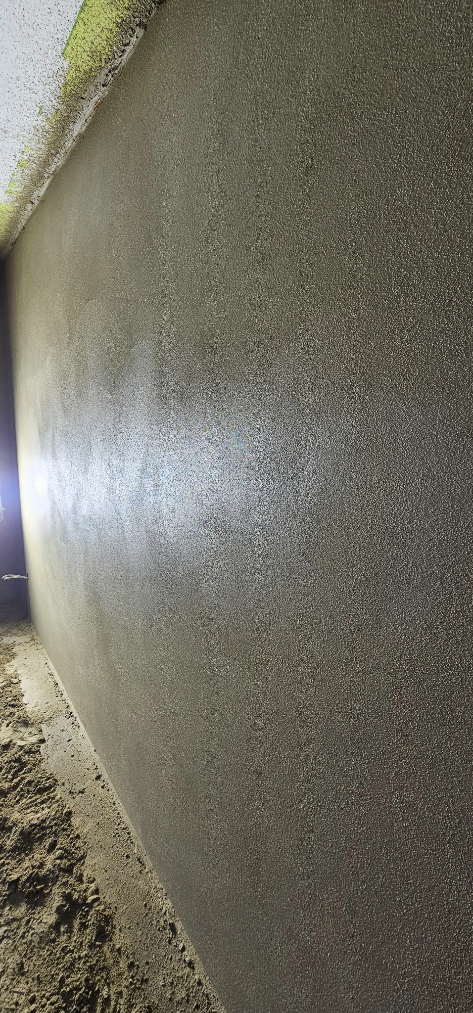 Świeżo nałożony, mokry tynk na ścianie wewnętrznej, z widocznymi śladami po narzędziach i piaskiem na podłodze. Delikatne światło podkreśla fakturę.