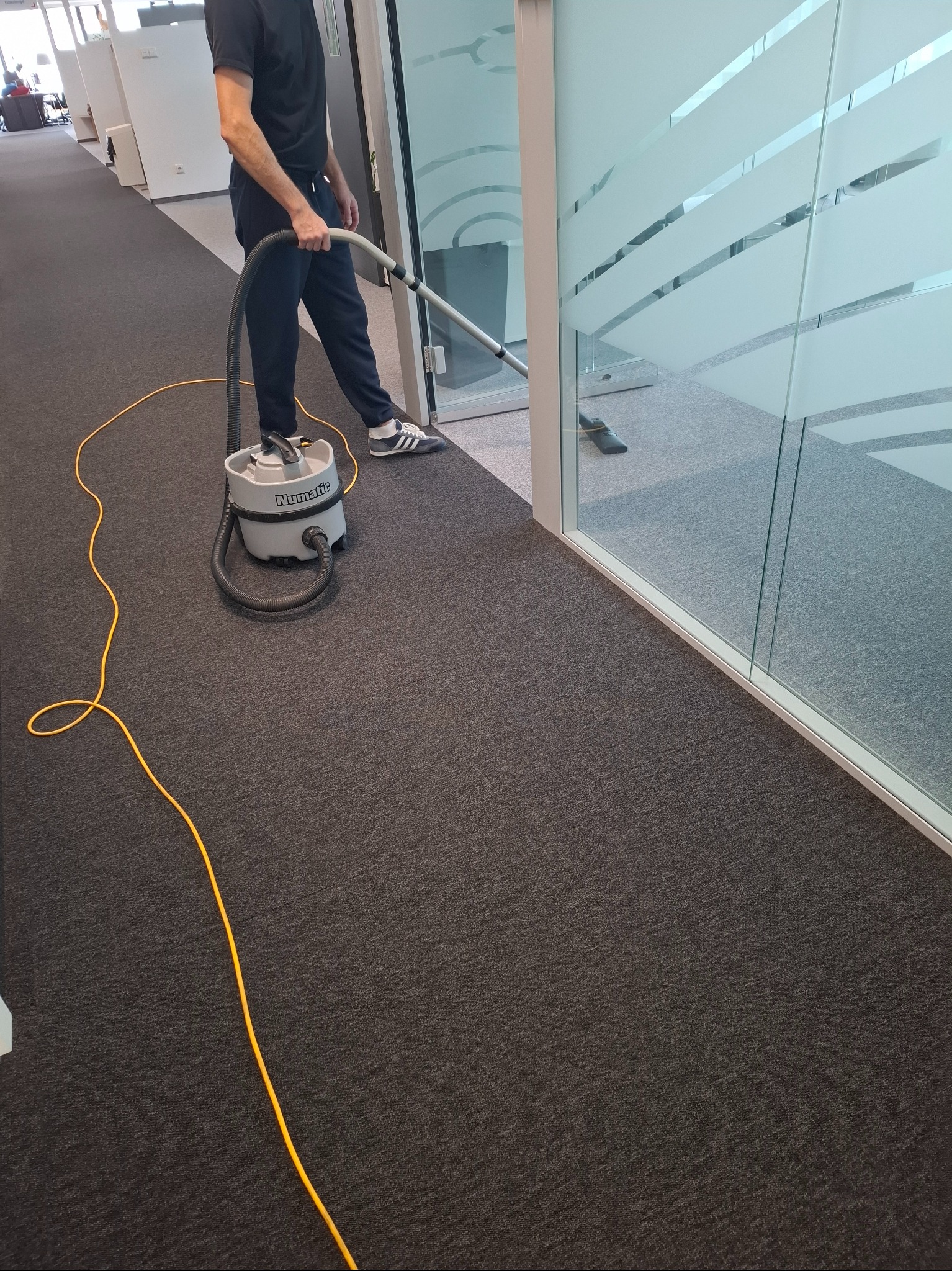 Osoba odkurza szary dywan w biurze za pomocą odkurzacza Numatic. Widoczne przeszklone biuro z matowymi pasami. Żółty kabel odkurzacza na podłodze.