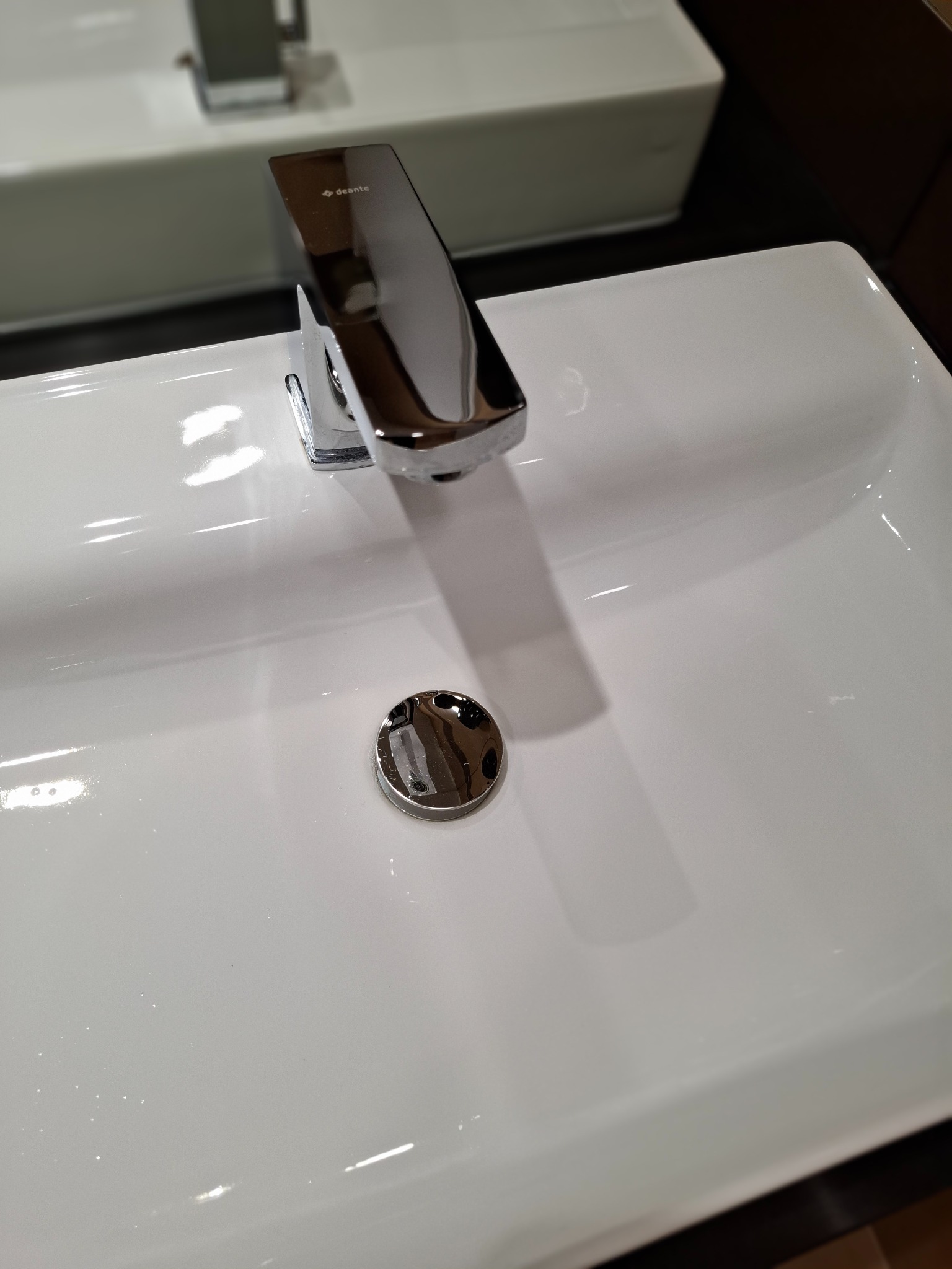 Błyszcząca, chromowana bateria umywalkowa Deante na czystej, białej umywalce o prostokątnym kształcie. Widoczny odpływ. Minimalistyczny design.