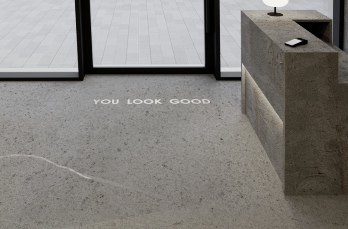 Minimalistyczne wnętrze z betonową recepcją i napisem 'YOU LOOK GOOD' na podłodze. Nowoczesny design z subtelnym oświetleniem LED.