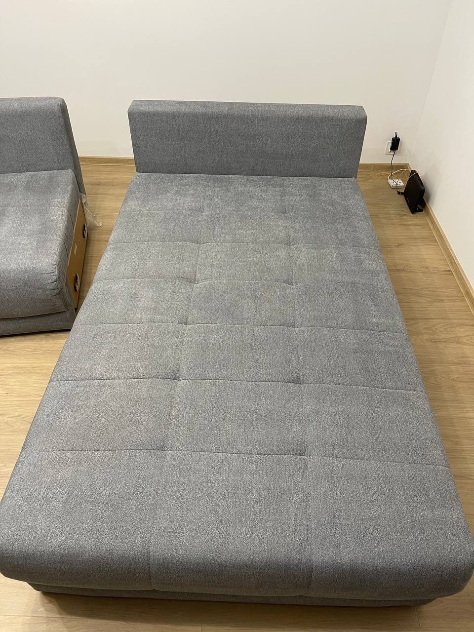 Rozłożona szara kanapa z tapicerowanymi segmentami, widoczna część sofy z boku. Minimalistyczne wnętrze, drewniana podłoga.