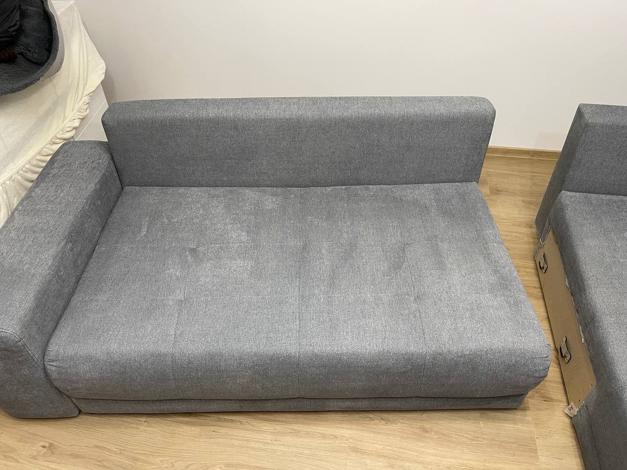 Rozłożona szara kanapa modułowa na drewnianej podłodze, ustawiona na tle białej ściany. Widoczny fragment narożnika. Gotowa do użytku po czyszczeniu.