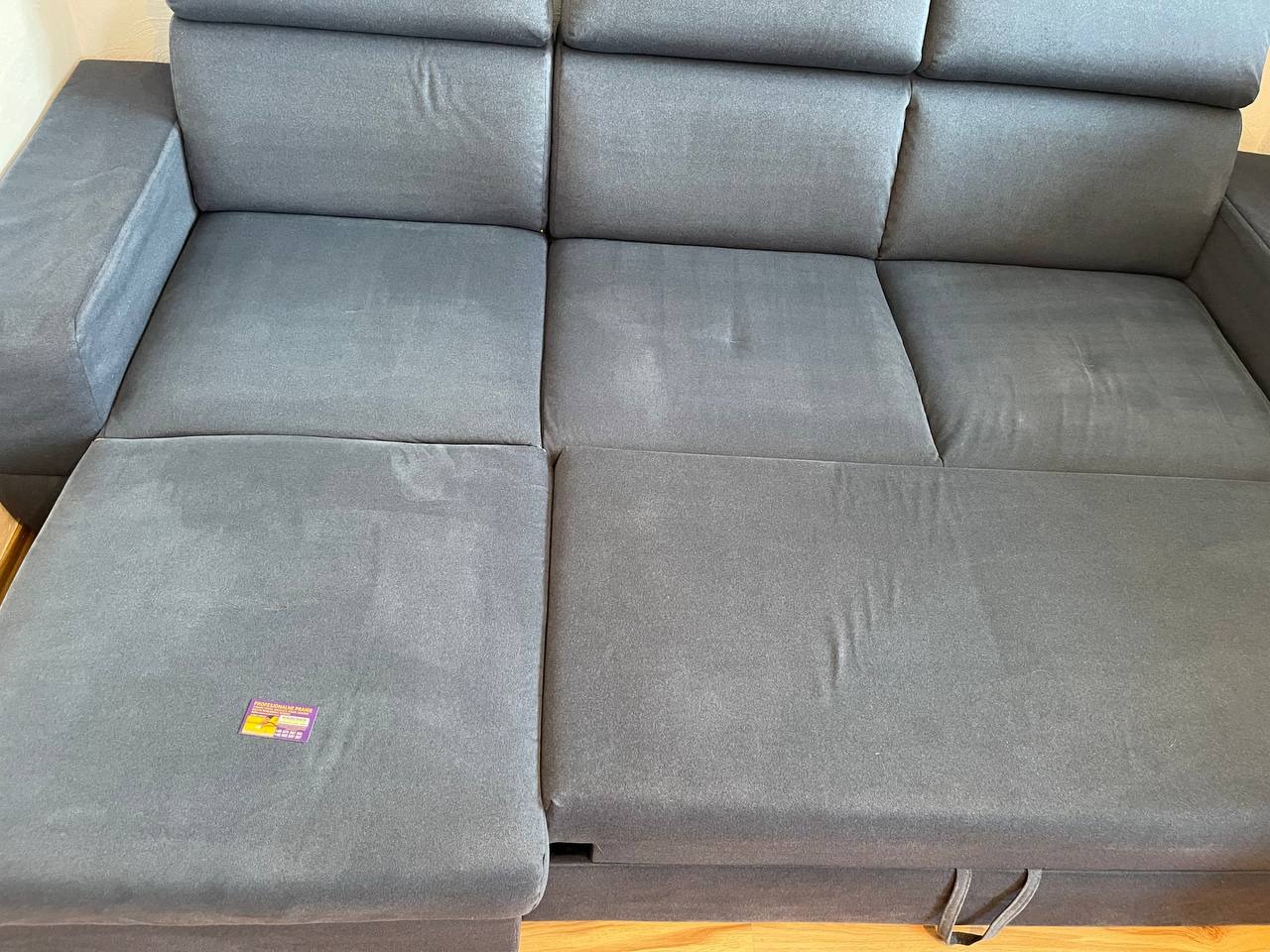 Rozłożona, szara kanapa z widoczną metką na jednym z siedzisk. Widoczne ślady użytkowania na tapicerce. Ujęcie z góry, w dziennym świetle.