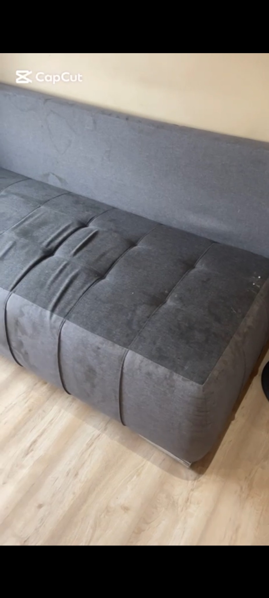 Szara, tapicerowana sofa z wyraźnymi śladami użytkowania, widocznymi zabrudzeniami na siedzisku i oparciu. Sofa stoi na jasnej, drewnianej podłodze.