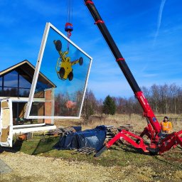 Mat-okna serwis pogwarancyjny okien - Montaż okna z użyciem dźwigu przyssawkowego na budowie domu jednorodzinnego. Widoczny pracownik w pomarańczowej kamizelce i fragment białego busa.