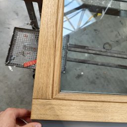 Mat-okna serwis pogwarancyjny okien - Fragment drewnianego okna z szybą, trzymanego przez dłoń, ukazujący detale wykończenia i połączenie z szarą listwą. Widoczne narzędzia na stole warsztatowym.