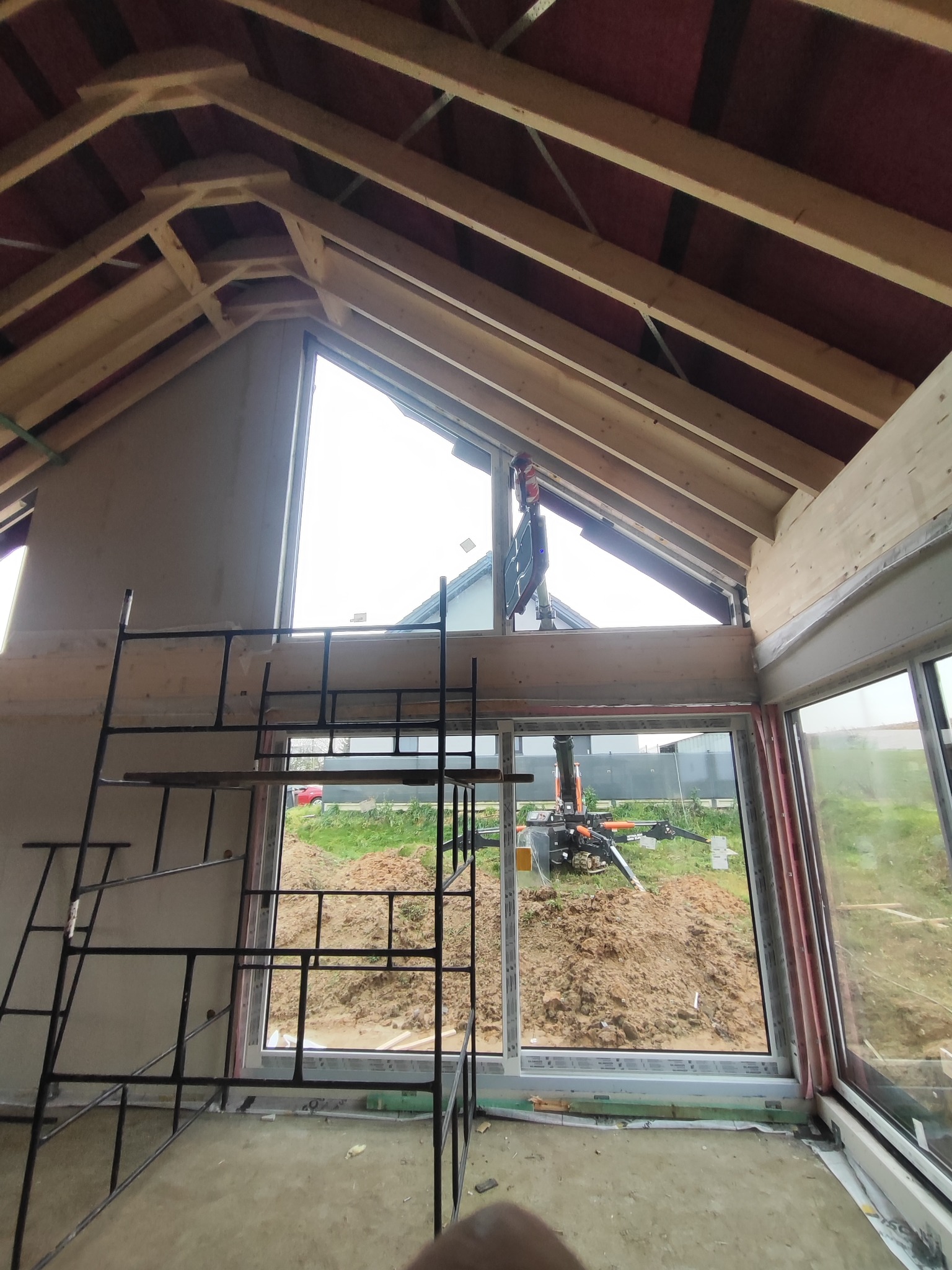 Montaż okna panoramicznego w surowym stanie domu z widoczną konstrukcją dachu i rusztowaniem. Na zewnątrz widoczny sprzęt budowlany.