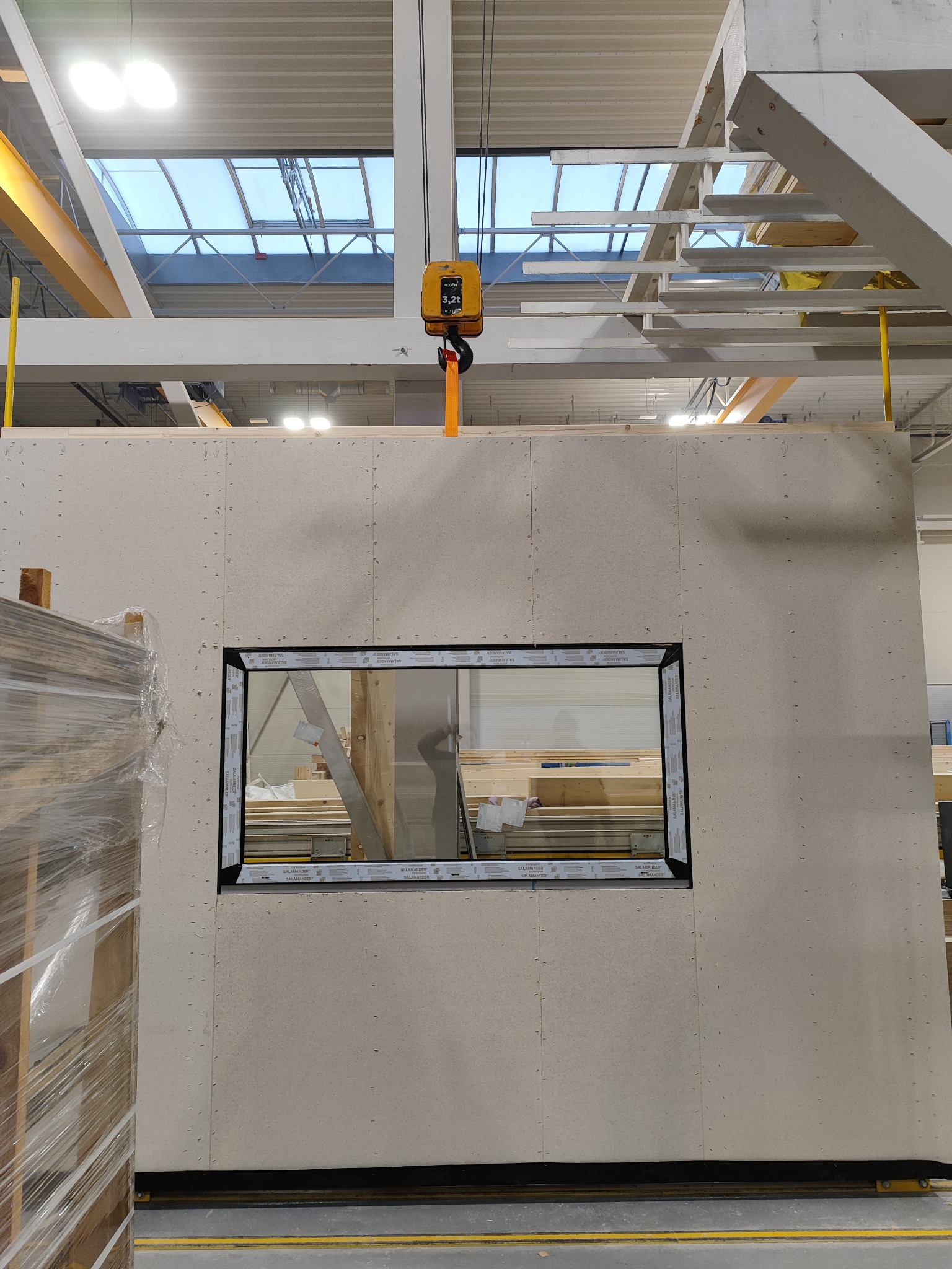 Wstawione okno PCV Salamander w prefabrykowanej ścianie z płyty OSB. Widoczny sufit hali z oknami dachowymi i suwnica. Paleta z materiałami budowlanymi po lewej.