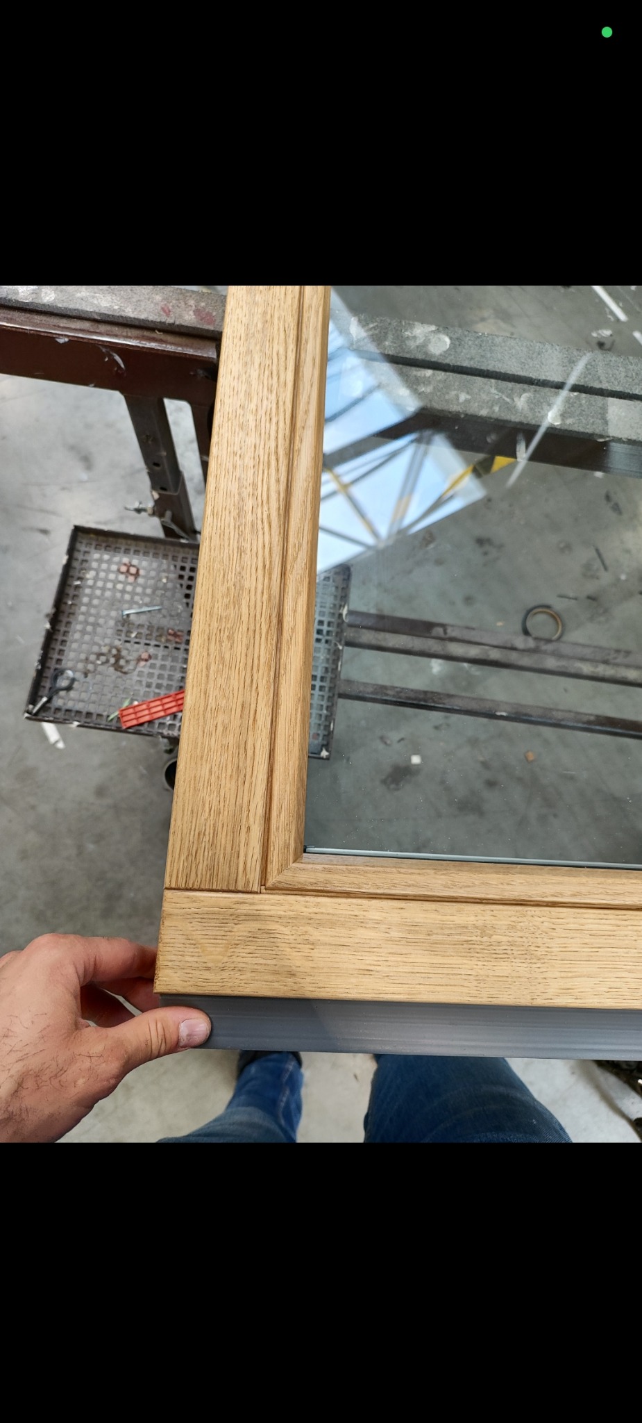 Fragment drewnianego okna z szybą, trzymanego przez dłoń, ukazujący detale wykończenia i połączenie z szarą listwą. Widoczne narzędzia na stole warsztatowym.