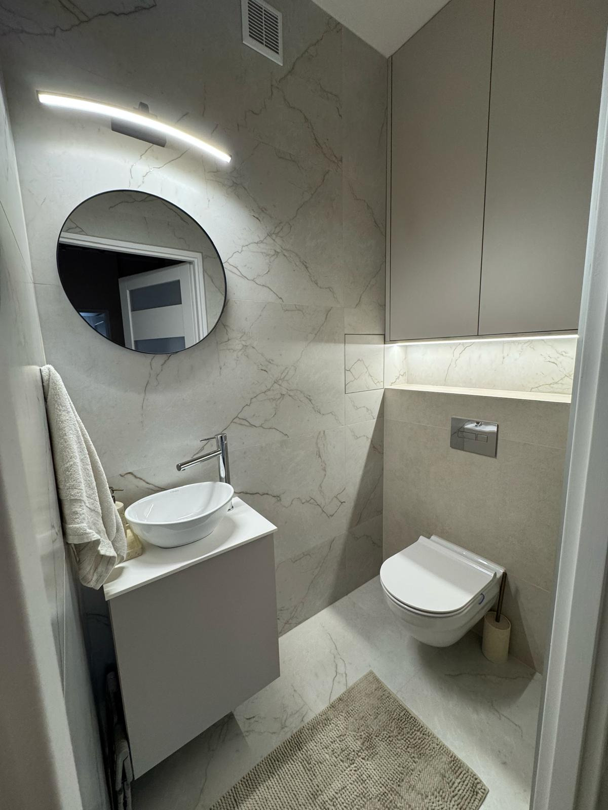 Nowoczesna łazienka z umywalką nablatową, owalnym lustrem z podświetleniem LED, wiszącą toaletą i szafkami w zabudowie. Marmurowe płytki na ścianach i podłodze.