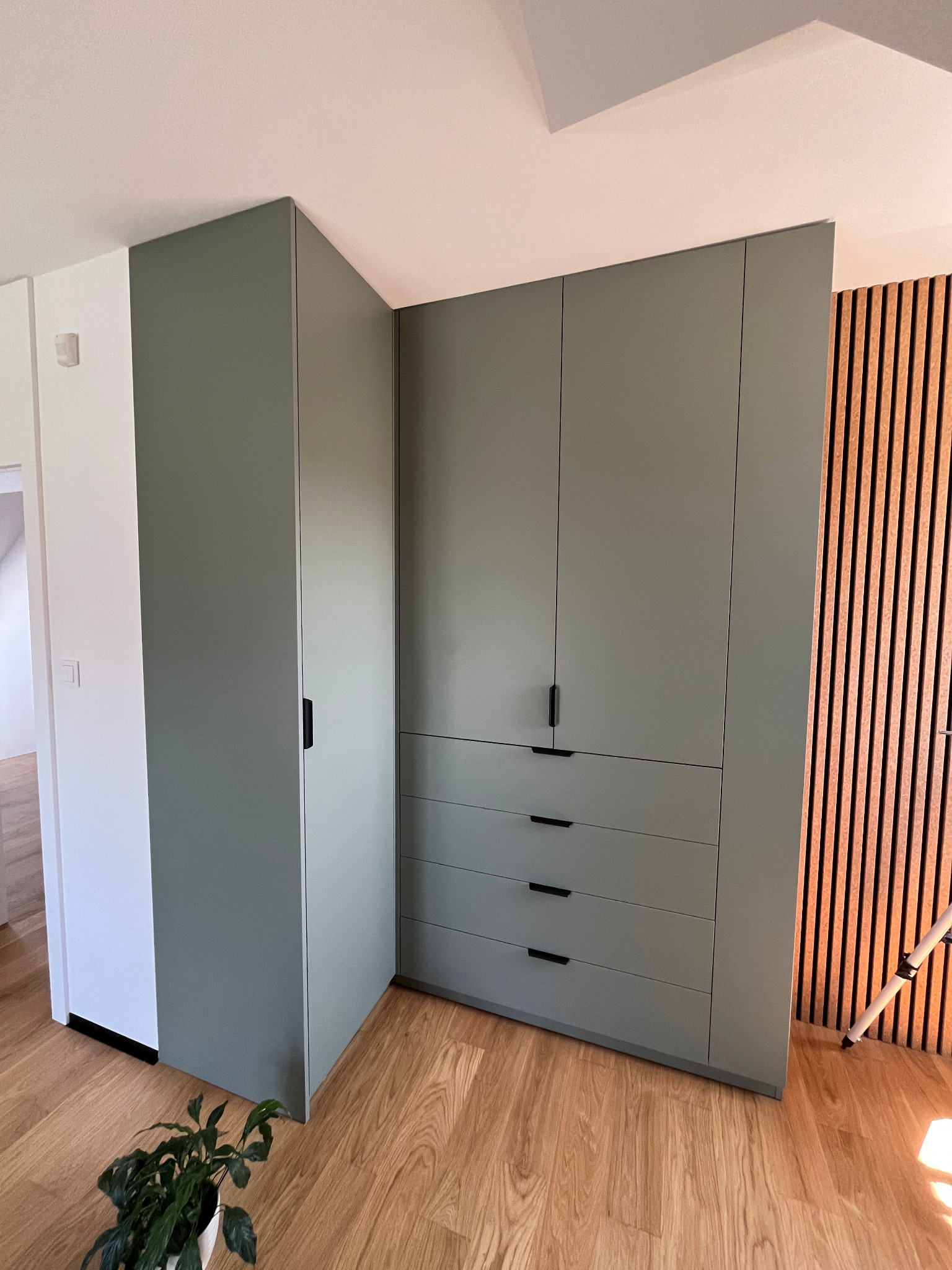 Zabudowa wnęki z szafą i szufladami w odcieniach zieleni, minimalistyczne uchwyty, drewniana podłoga i dekoracyjna ściana z lameli w tle. Nowoczesny design.