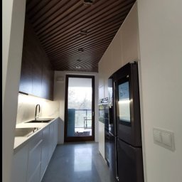 Reda meble - Nowoczesna kuchnia z drewnianym sufitem lamelowym i białymi szafkami, widok na balkon. Minimalistyczny design, eleganckie wykończenie, czarne AGD.