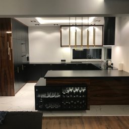 Reda meble - Nowoczesna kuchnia z wyspą: czarne szafki, drewniane akcenty, marmurowe podłogi i złote lampy wiszące. Widoczna przeszklona szafka z kieliszkami.