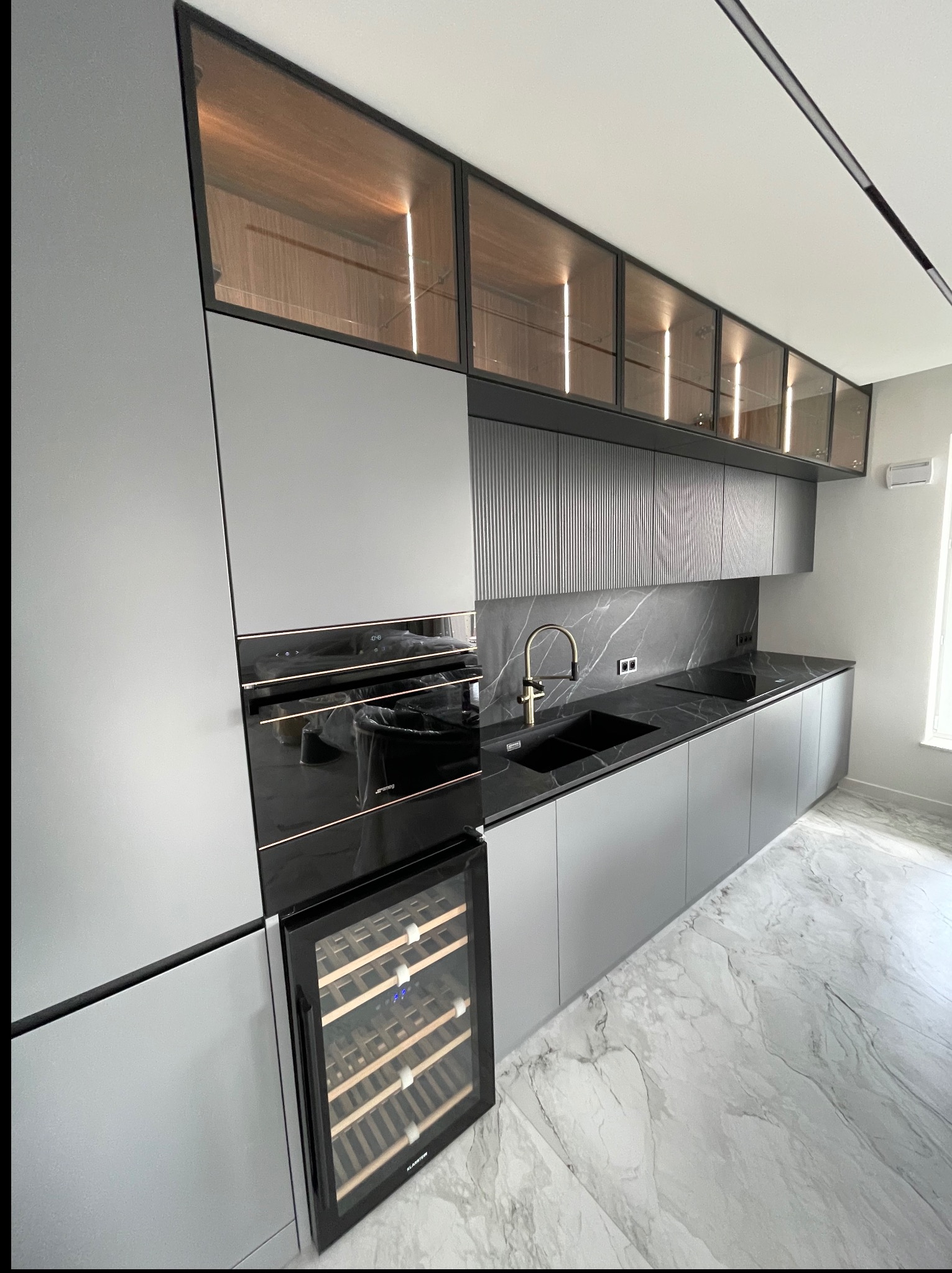 Nowoczesna kuchnia w odcieniach szarości z czarnym blatem imitującym marmur, złotą armaturą i przeszklonymi szafkami górnymi z oświetleniem LED.