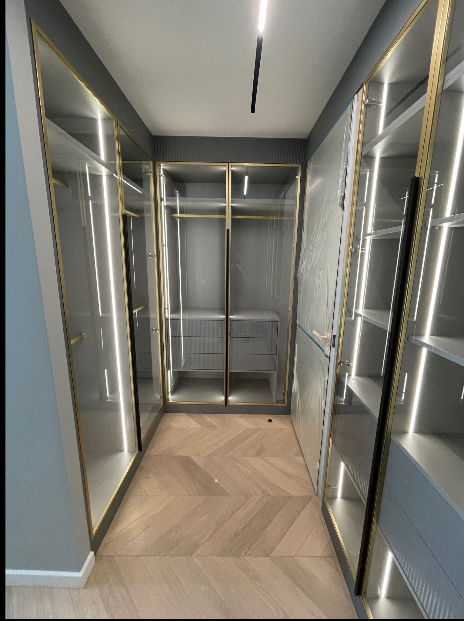 Nowoczesna garderoba z podświetlanymi półkami i złotymi ramami, parkiet w jodełkę. Część szafy jest jeszcze zabezpieczona folią ochronną.