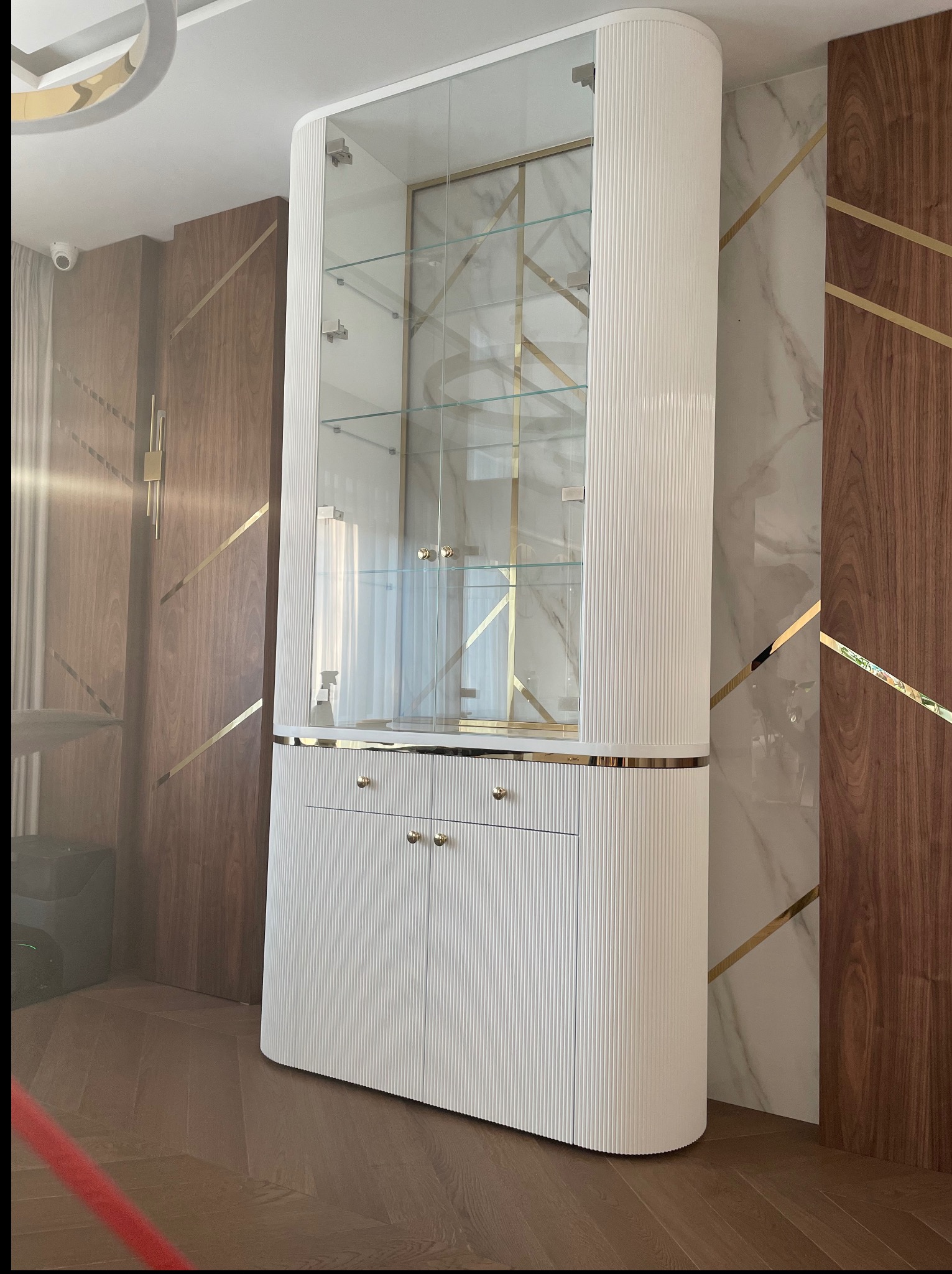 Biała, ryflowana witryna narożna z przeszkloną górą i złotymi akcentami, wkomponowana w eleganckie wnętrze z drewnianymi i marmurowymi panelami.