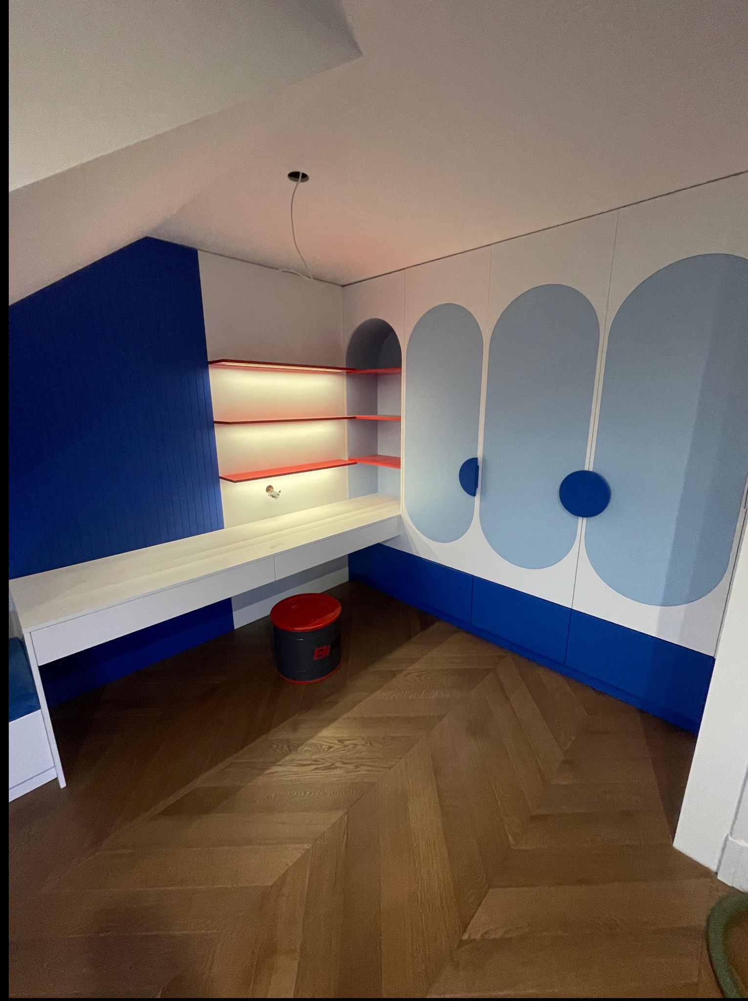 Biało-niebieska szafa wnękowa z biurkiem i półkami podświetlonymi na czerwono, w pokoju z parkietem ułożonym w jodełkę. Nowoczesny design, oświetlenie LED.