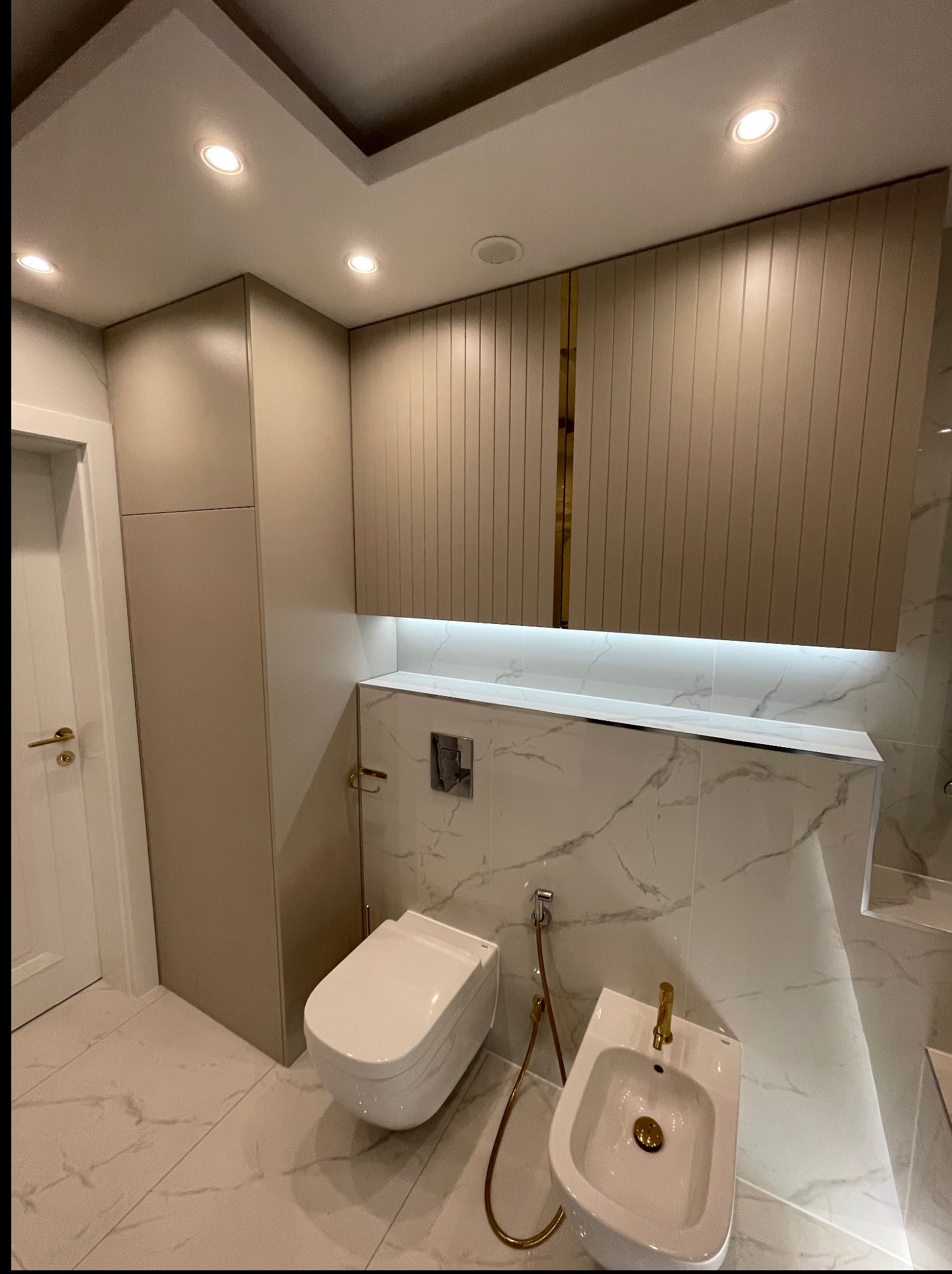 Elegancka łazienka z zabudową meblową w odcieniach beżu i bieli, marmurowe płytki, podwieszana toaleta i bidet ze złotymi detalami, oświetlenie LED.