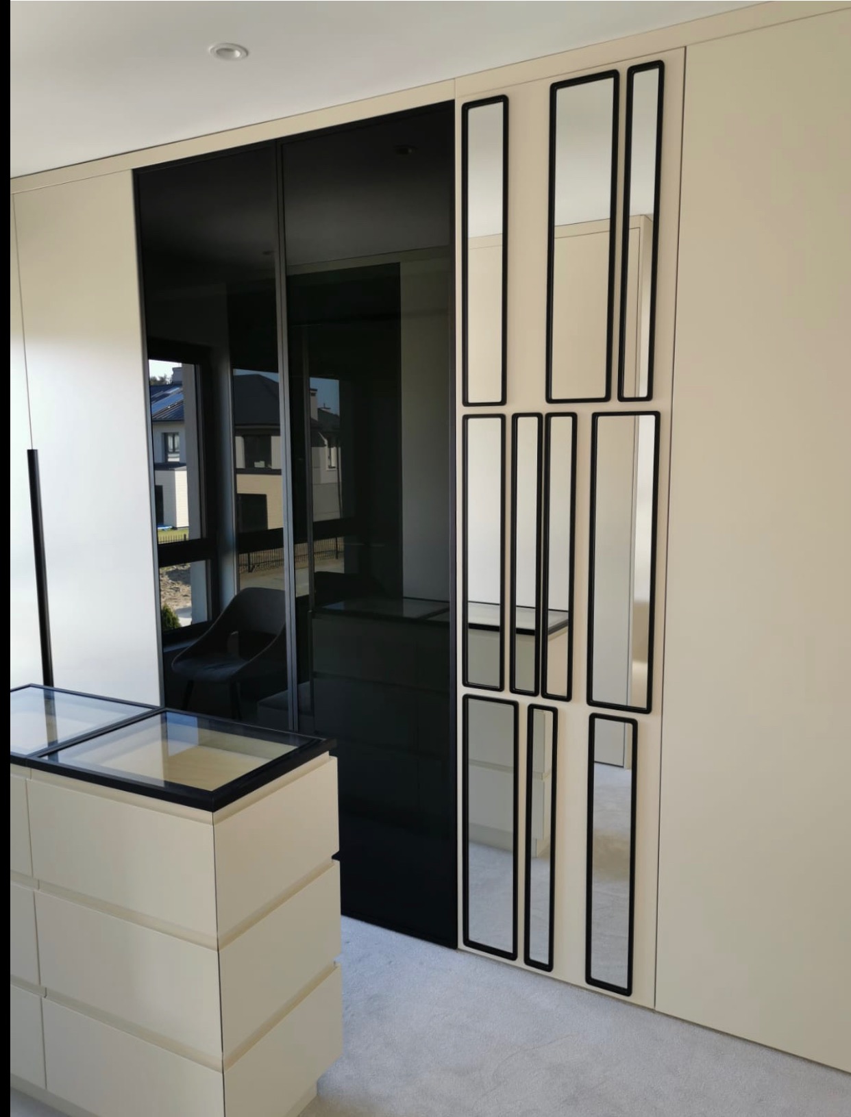 Nowoczesna szafa wnękowa z czarnymi, lustrzanymi drzwiami i zdobieniami z prostokątnych luster. Beżowe korpusy, szklana komoda z szufladami.