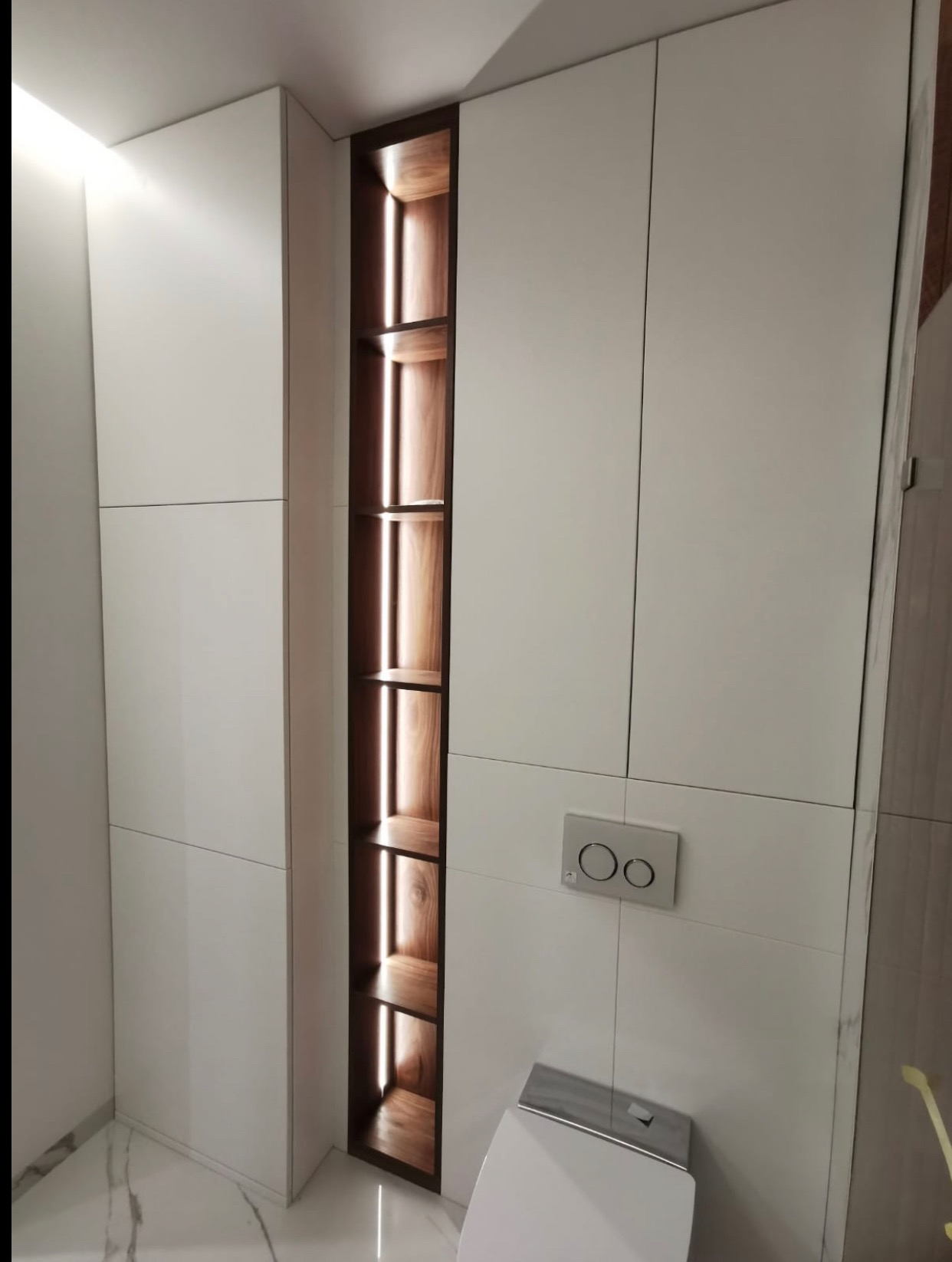 Biała szafa wnękowa z podświetlanymi półkami z drewna w nowoczesnej łazience z marmurową podłogą i płytkami. Minimalistyczny design i funkcjonalność.