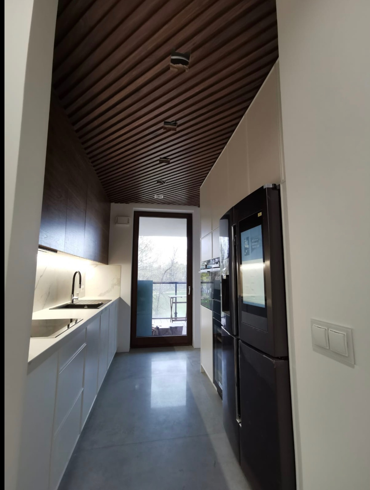 Nowoczesna kuchnia z drewnianym sufitem lamelowym i białymi szafkami, widok na balkon. Minimalistyczny design, eleganckie wykończenie, czarne AGD.