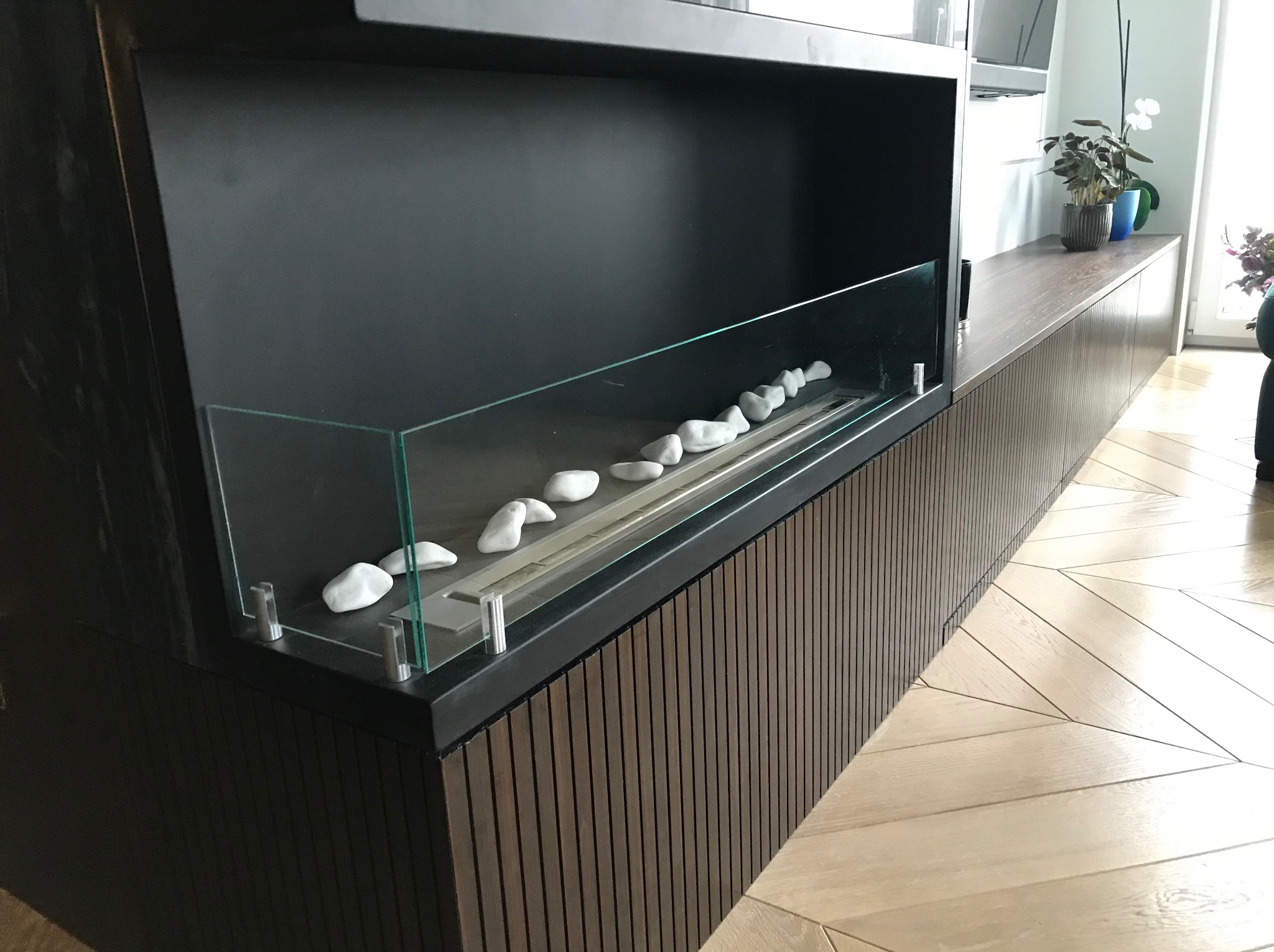 Ciemna szafka RTV z biokominkiem, zabezpieczonym szkłem i białymi kamieniami, na tle parkietu w jodełkę i roślin doniczkowych. Nowoczesny design.