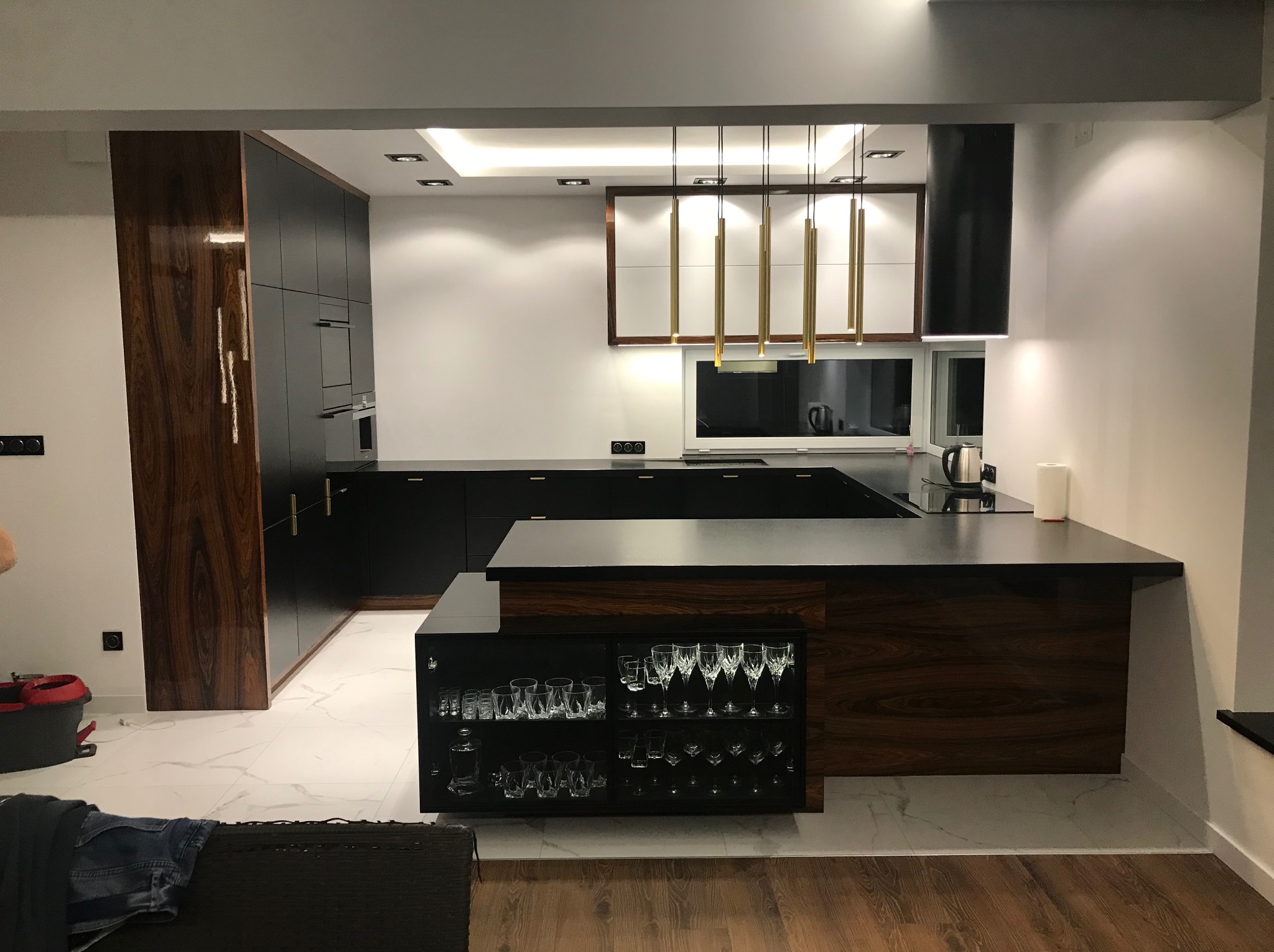 Nowoczesna kuchnia z wyspą: czarne szafki, drewniane akcenty, marmurowe podłogi i złote lampy wiszące. Widoczna przeszklona szafka z kieliszkami.