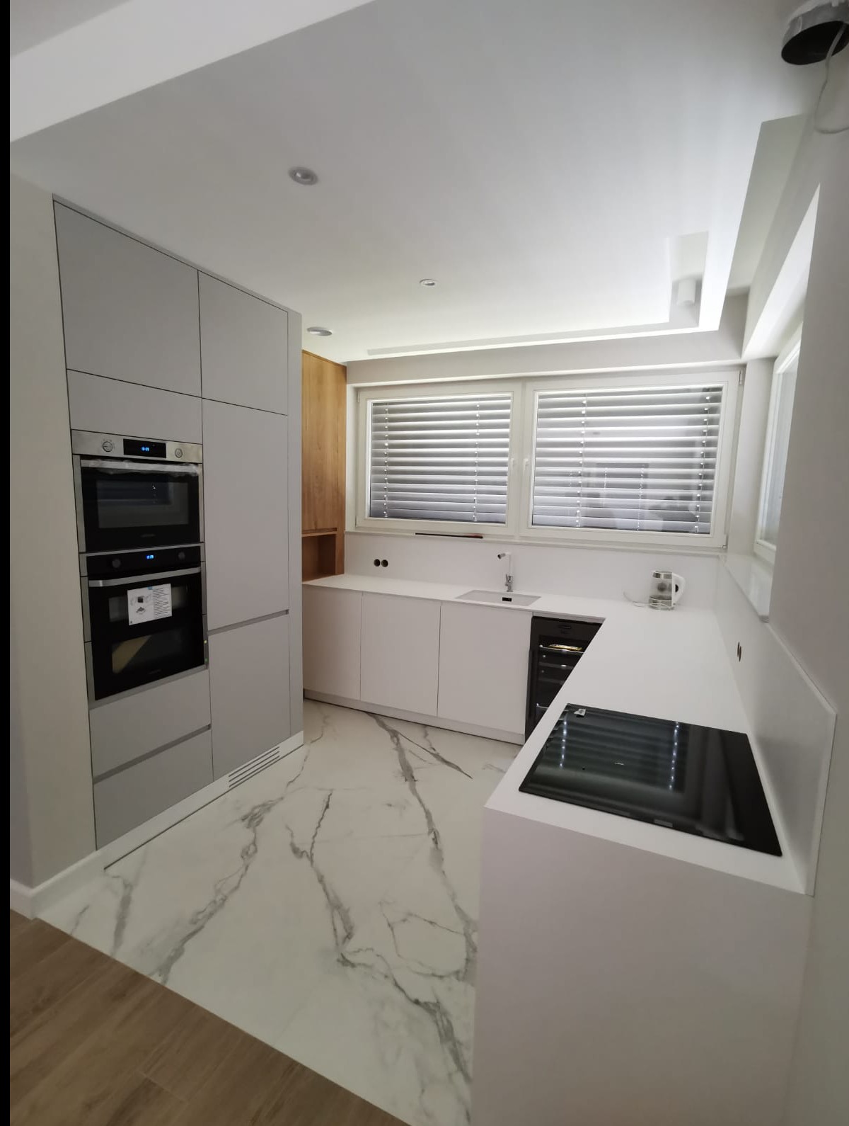 Nowoczesna kuchnia z szarymi szafkami i białym blatem, płyta indukcyjna, marmurowa podłoga, okno z żaluzjami, minimalistyczny design, oświetlenie punktowe.