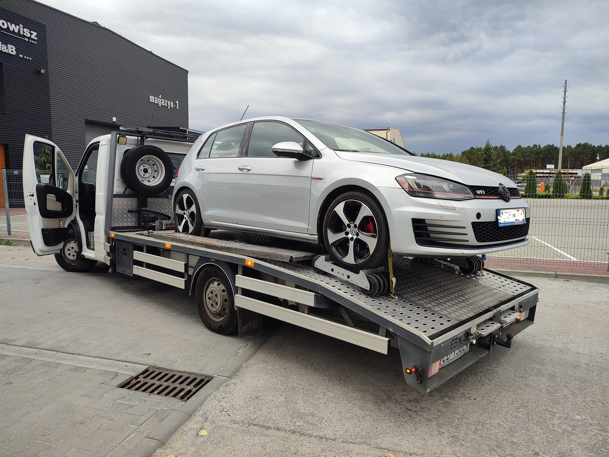 Srebrny Volkswagen Golf GTI na lawecie z otwartymi drzwiami kabiny, kołem zapasowym i budynkiem magazynu w tle. Usługa transportu pojazdu.