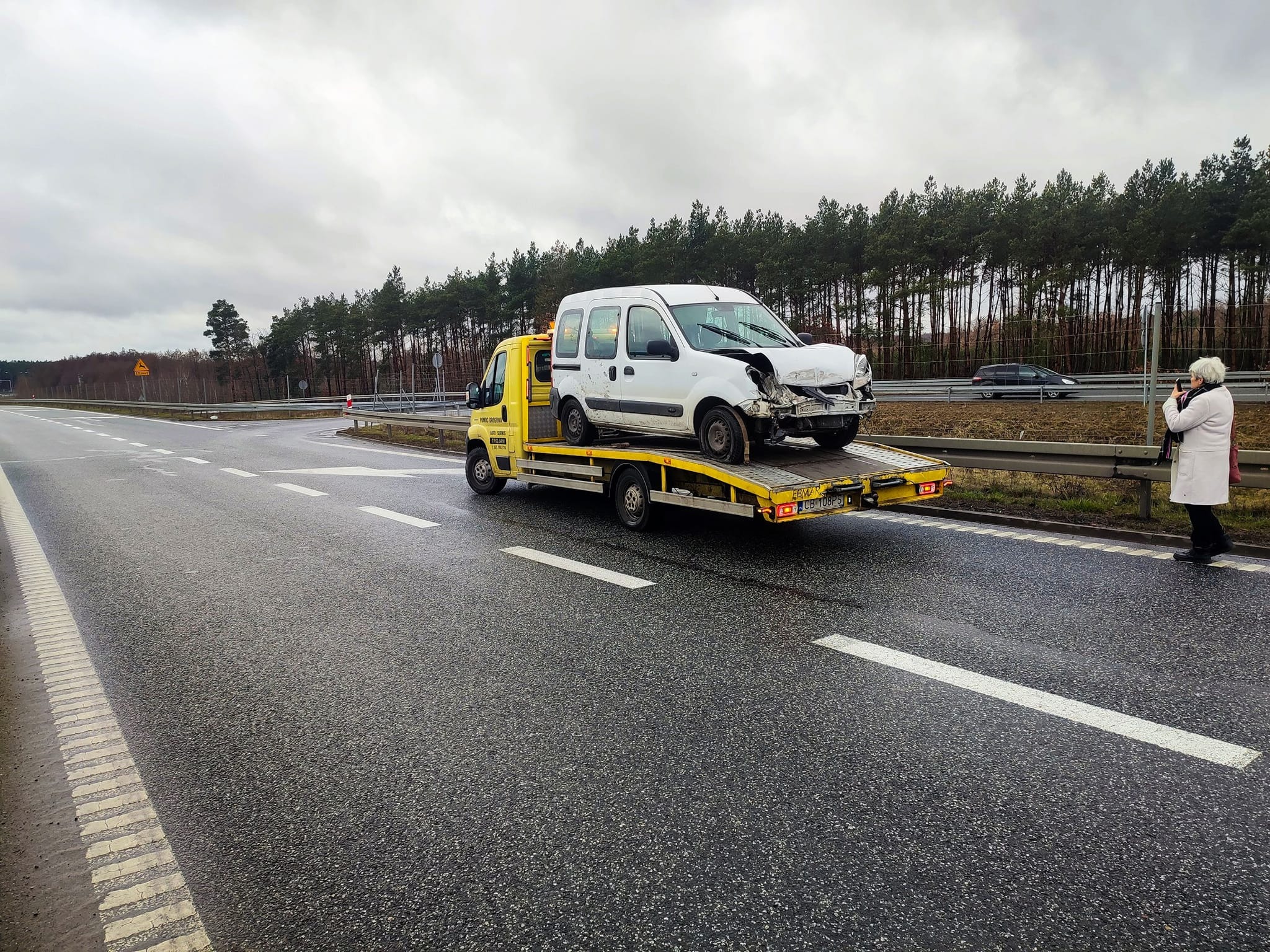 Żółta laweta z uszkodzonym białym vanem na platformie, stojąca na poboczu drogi. W tle las i osoba robiąca zdjęcie telefonem. Pochmurne niebo.