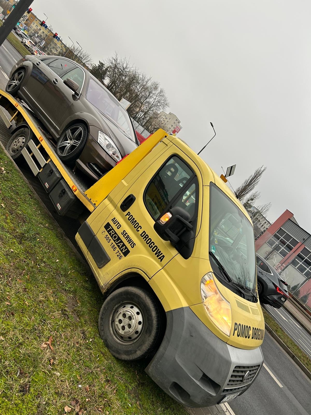Żółta laweta 'Pomoc Drogowa Trojan' transportuje brązowy samochód osobowy na tle miejskiego krajobrazu w pochmurny dzień. Widoczne budynki i sygnalizacja świetlna.