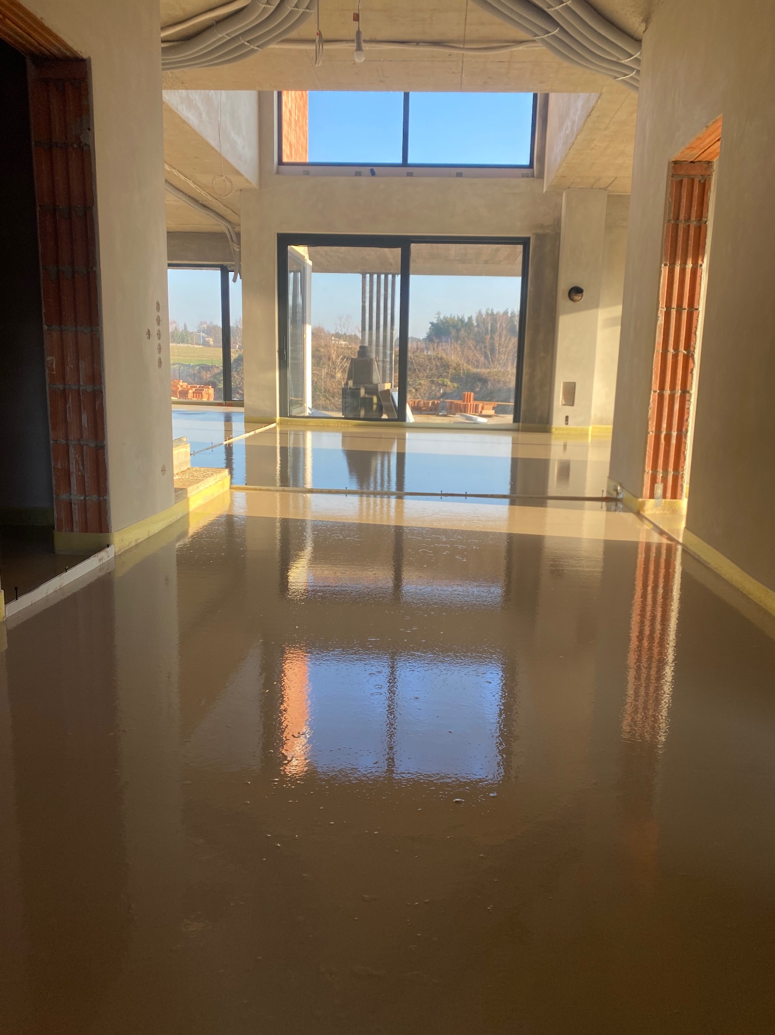 Świeżo wylana, gładka posadzka w surowym wnętrzu z dużymi oknami, odbijającymi niebo. Widoczne rury pod sufitem i ceglane elementy konstrukcyjne.