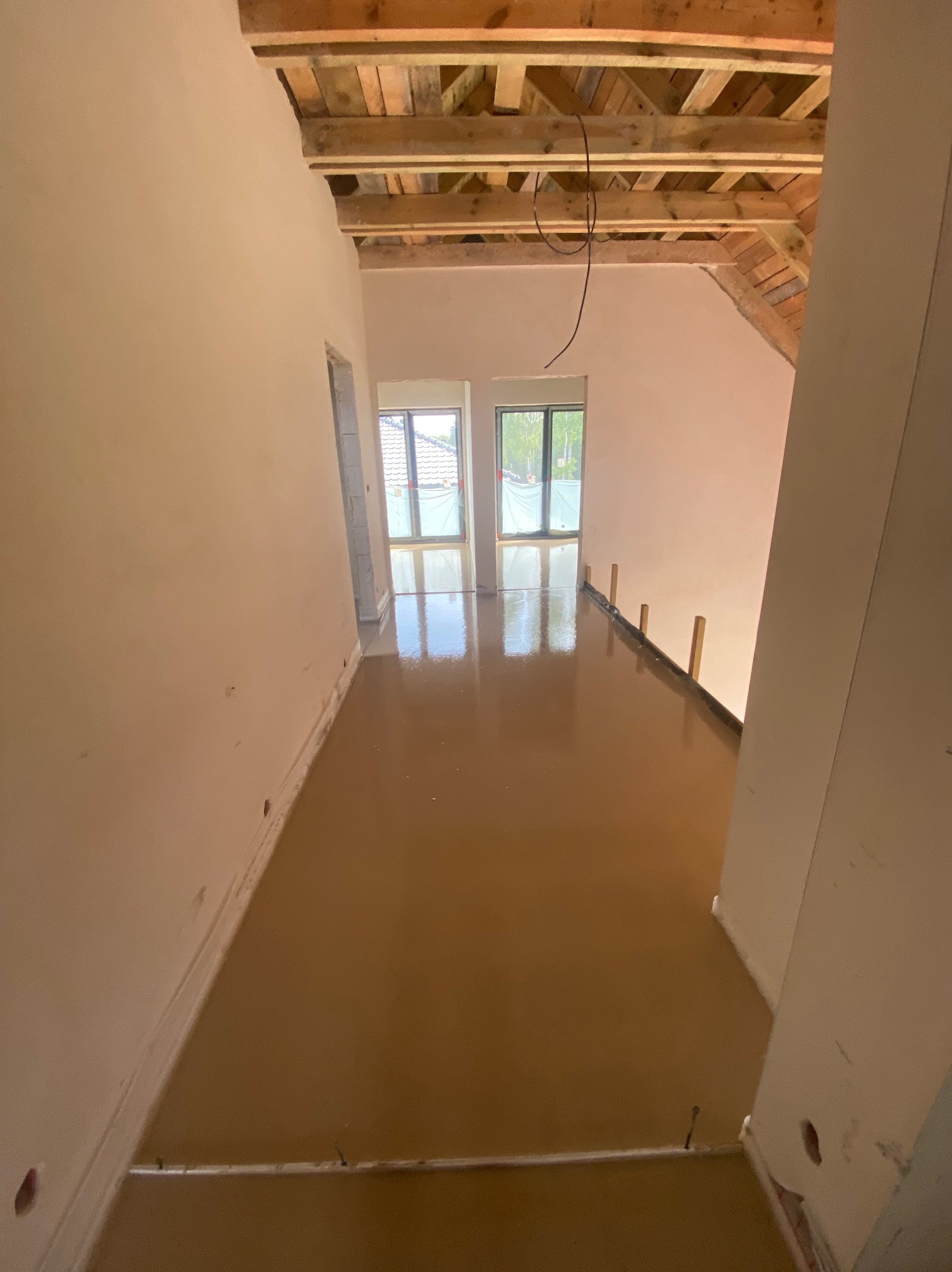 Świeżo wylana, brązowa posadzka w jasnym wnętrzu z drewnianym stropem. Widoczne okna i przygotowane listwy dylatacyjne. Podłoże odbija światło.