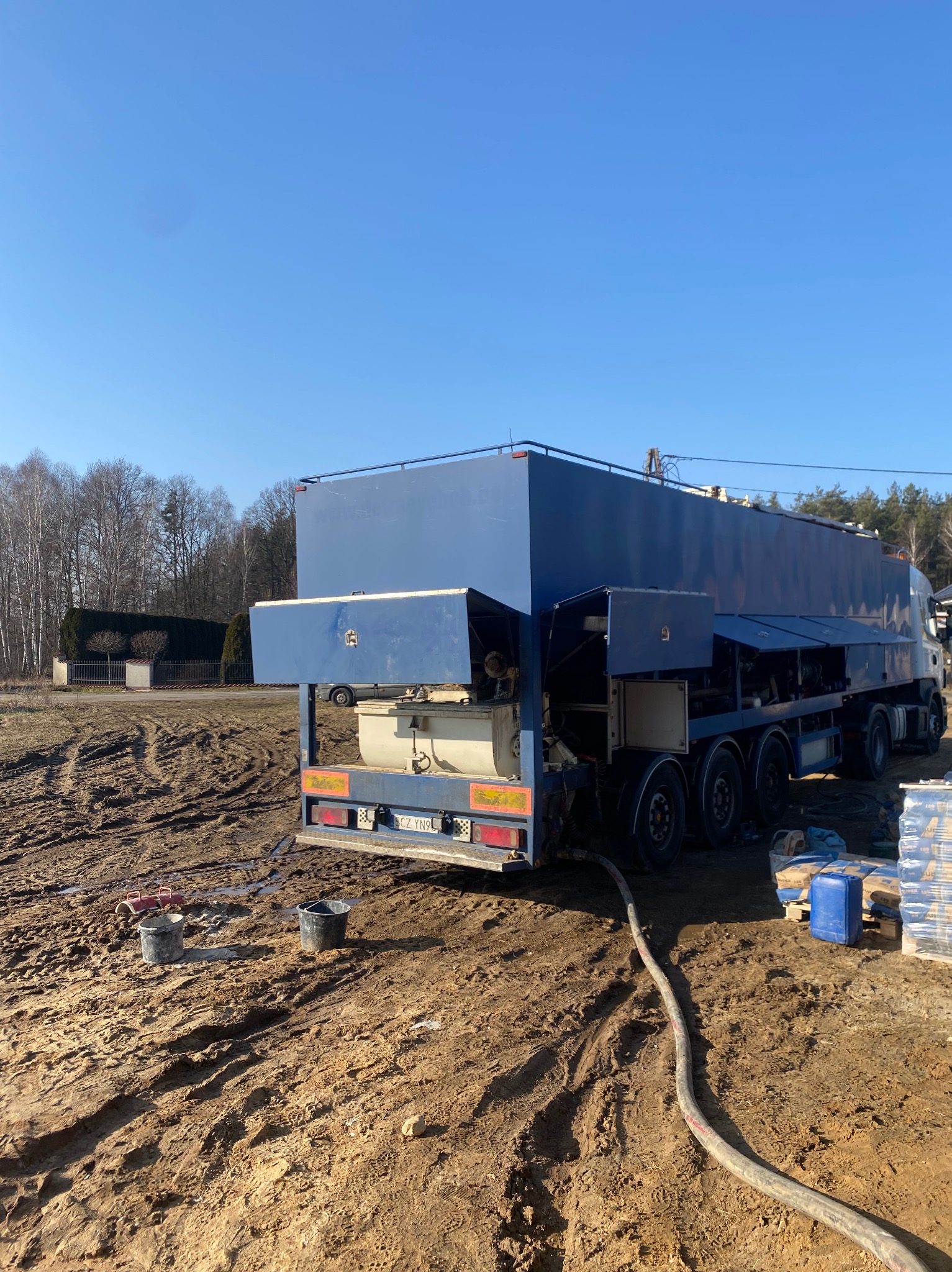 Niebieski samochód do wylewek stoi na błotnistym terenie. Wąż prowadzi do maszyny. Worki z materiałami budowlanymi obok. Błękitne niebo w tle.