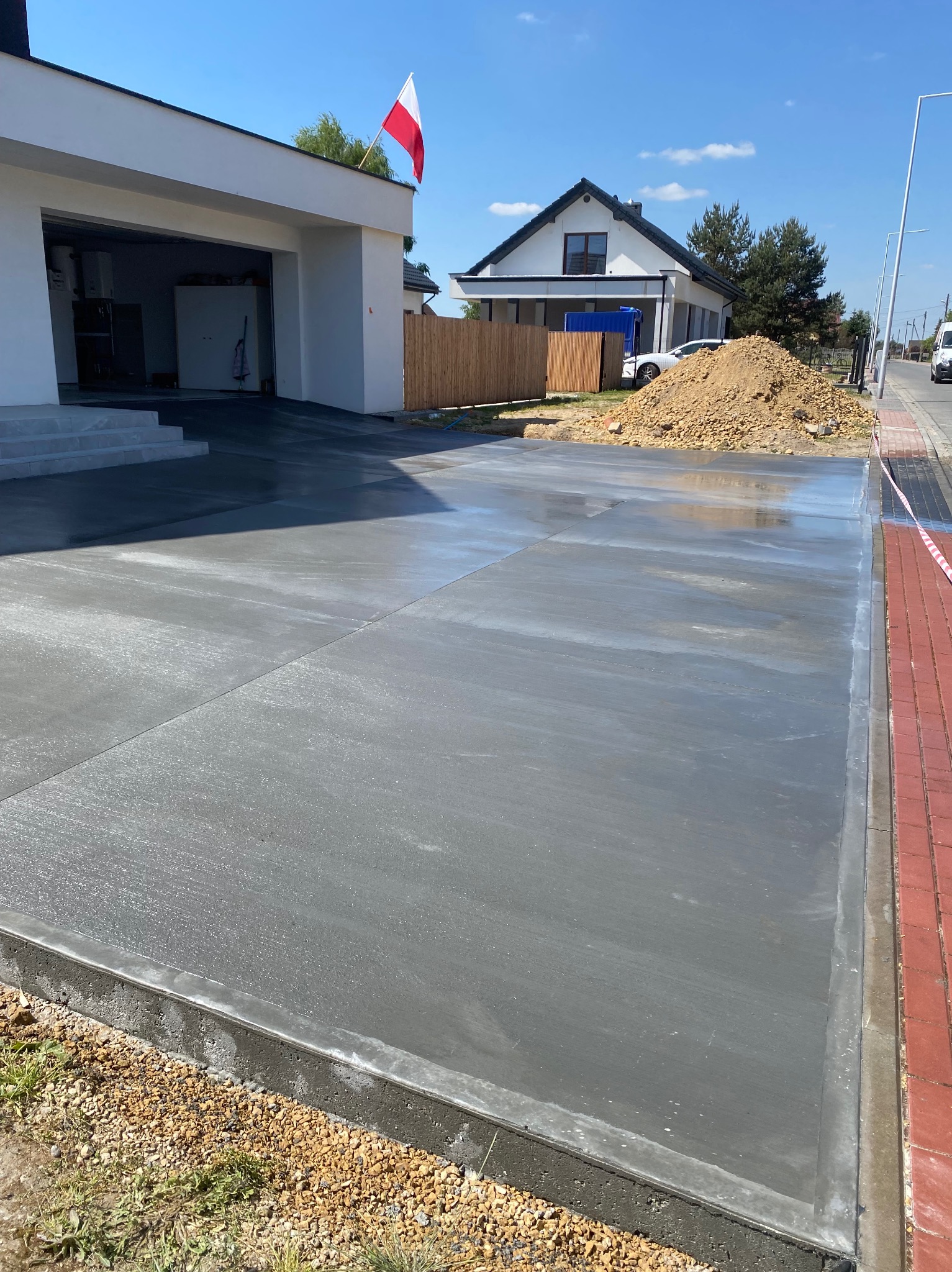 Świeżo wylana, gładka, szara posadzka na podjeździe domu jednorodzinnego w słoneczny dzień. W tle dom z polską flagą i sterta ziemi.