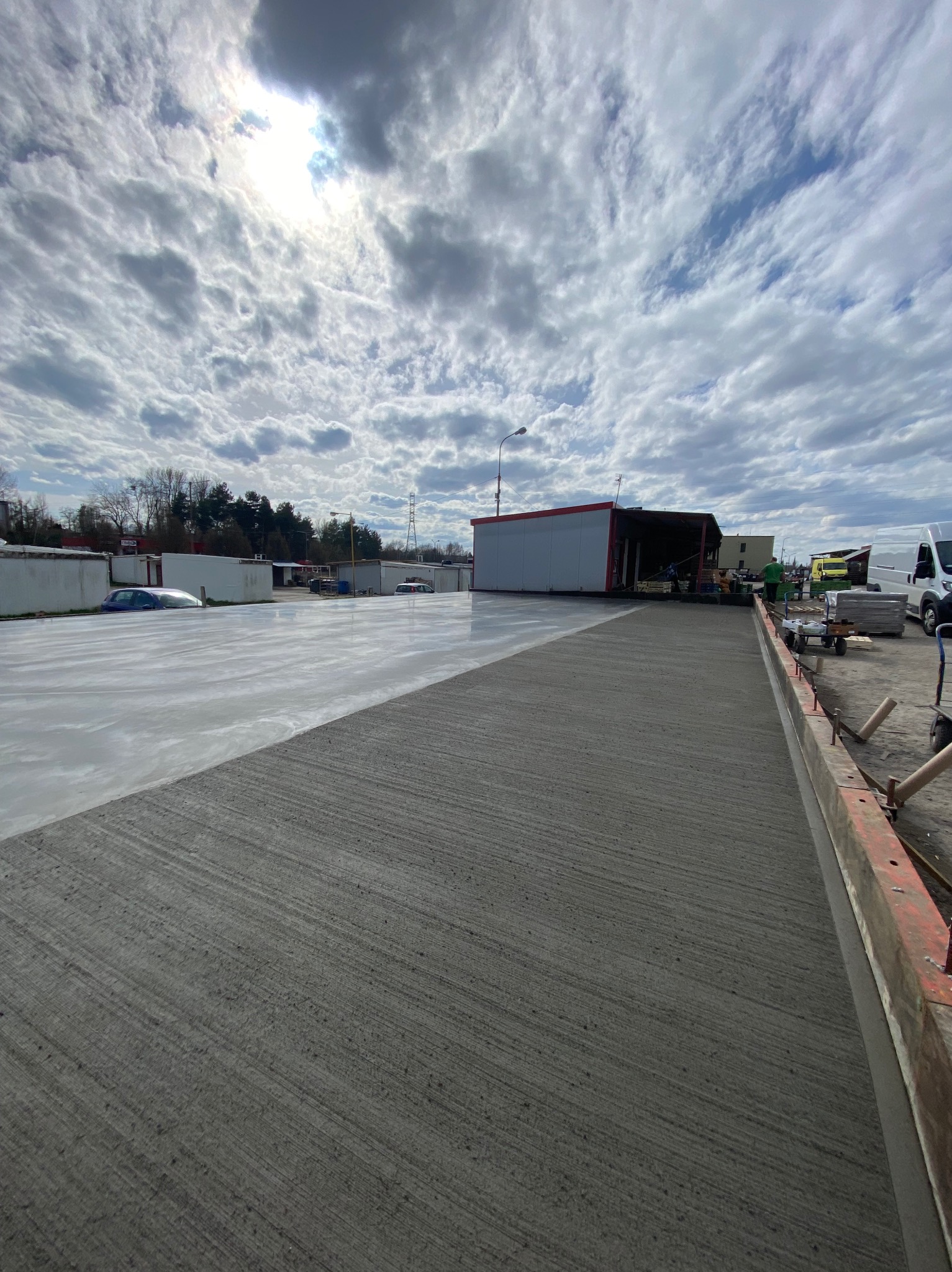 Świeżo wylana, gładka posadzka przemysłowa na zewnątrz budynku, częściowo mokra, z widocznymi narzędziami i ekipą w tle pod błękitnym niebem z chmurami.