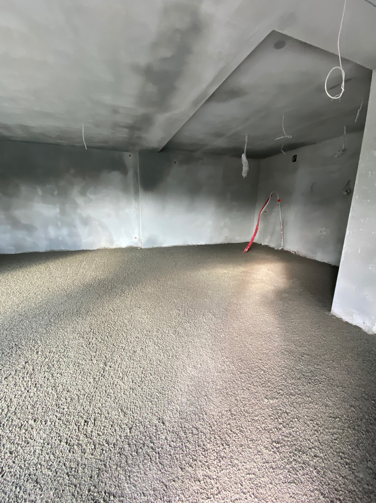 Wnętrze z wylaną, szarą posadzką, widoczne przewody elektryczne na ścianie i suficie. Świeża wylewka, surowy stan wykończenia, przygotowanie do dalszych prac.