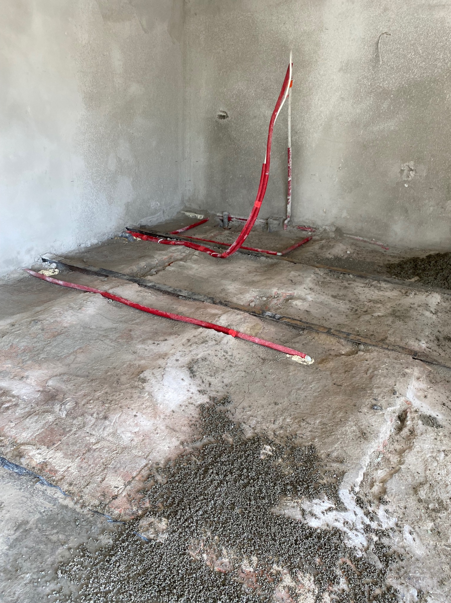 Przygotowanie do wylewki: czerwone rury ogrzewania podłogowego zamocowane na listwach, częściowo zalane wylewką. Surowe ściany w tle.