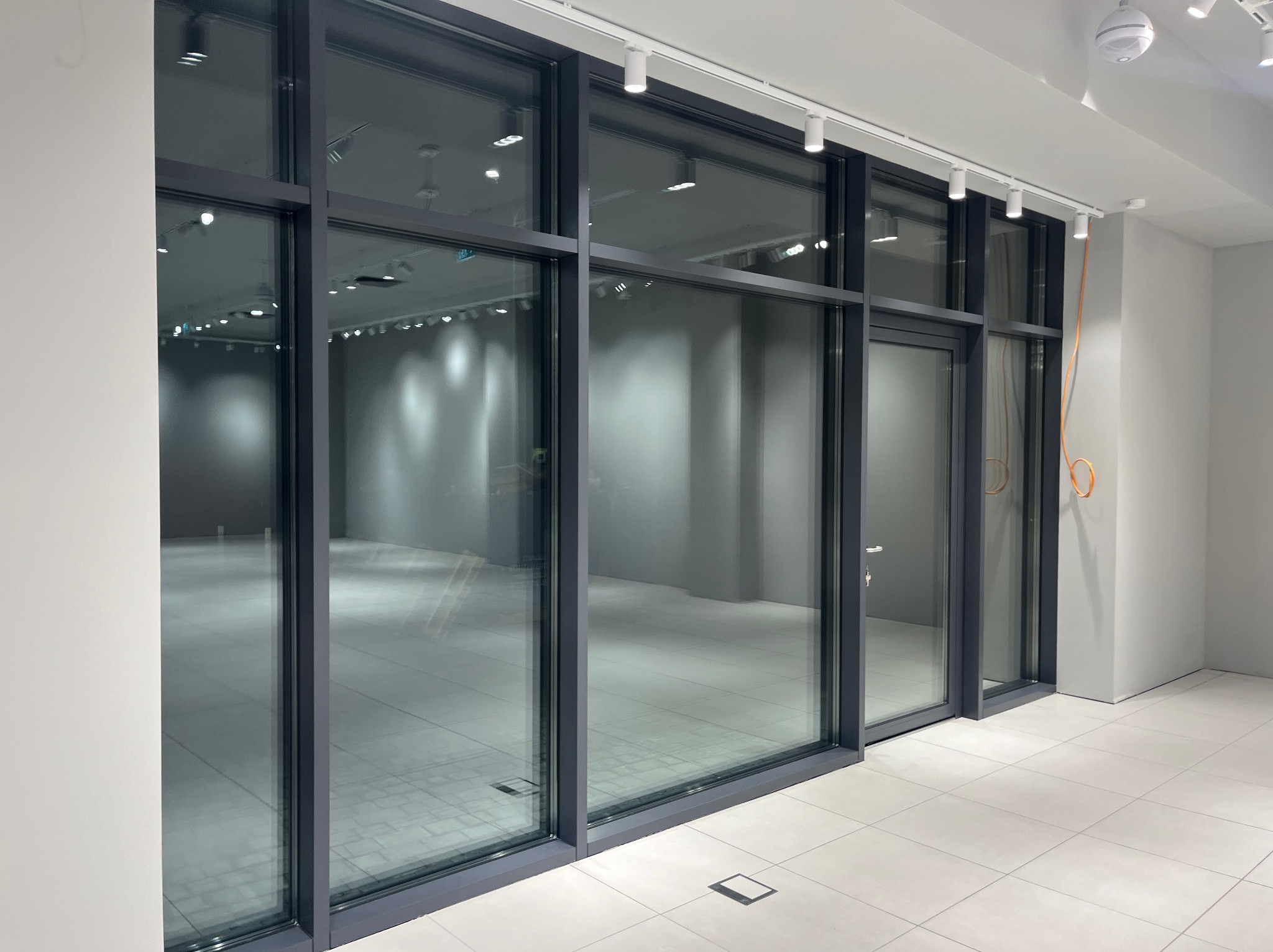 Puste, jasne wnętrze biurowe z dużymi oknami i szarą ramą. Widoczne białe płytki podłogowe i oświetlenie sufitowe. Minimalistyczny design, gotowe do sprzątania po remoncie.