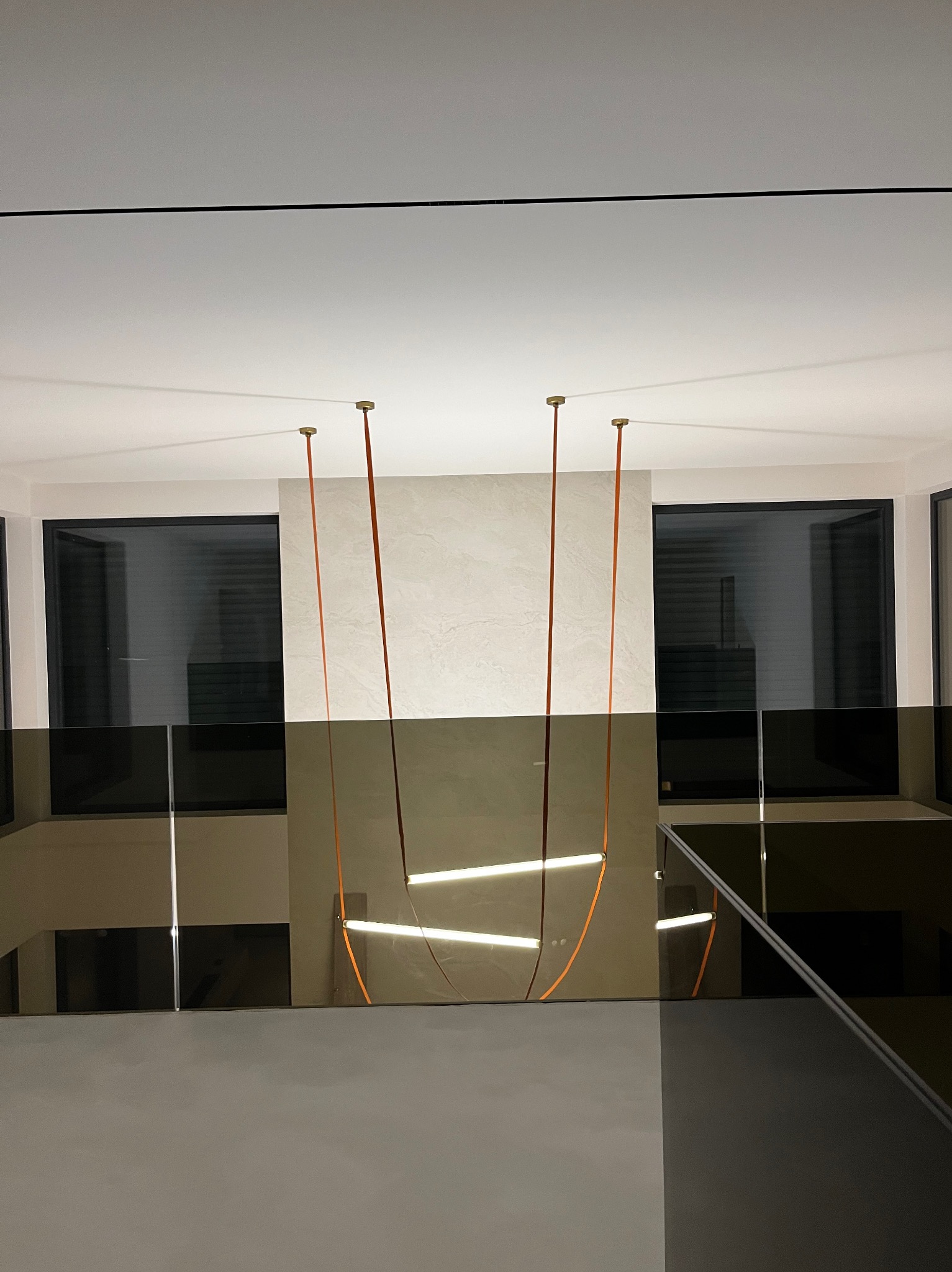 Nowoczesne wnętrze z minimalistyczną lampą wiszącą na tle okien i szklanej balustrady. Jasne ściany i stonowana kolorystyka podkreślają elegancję przestrzeni.