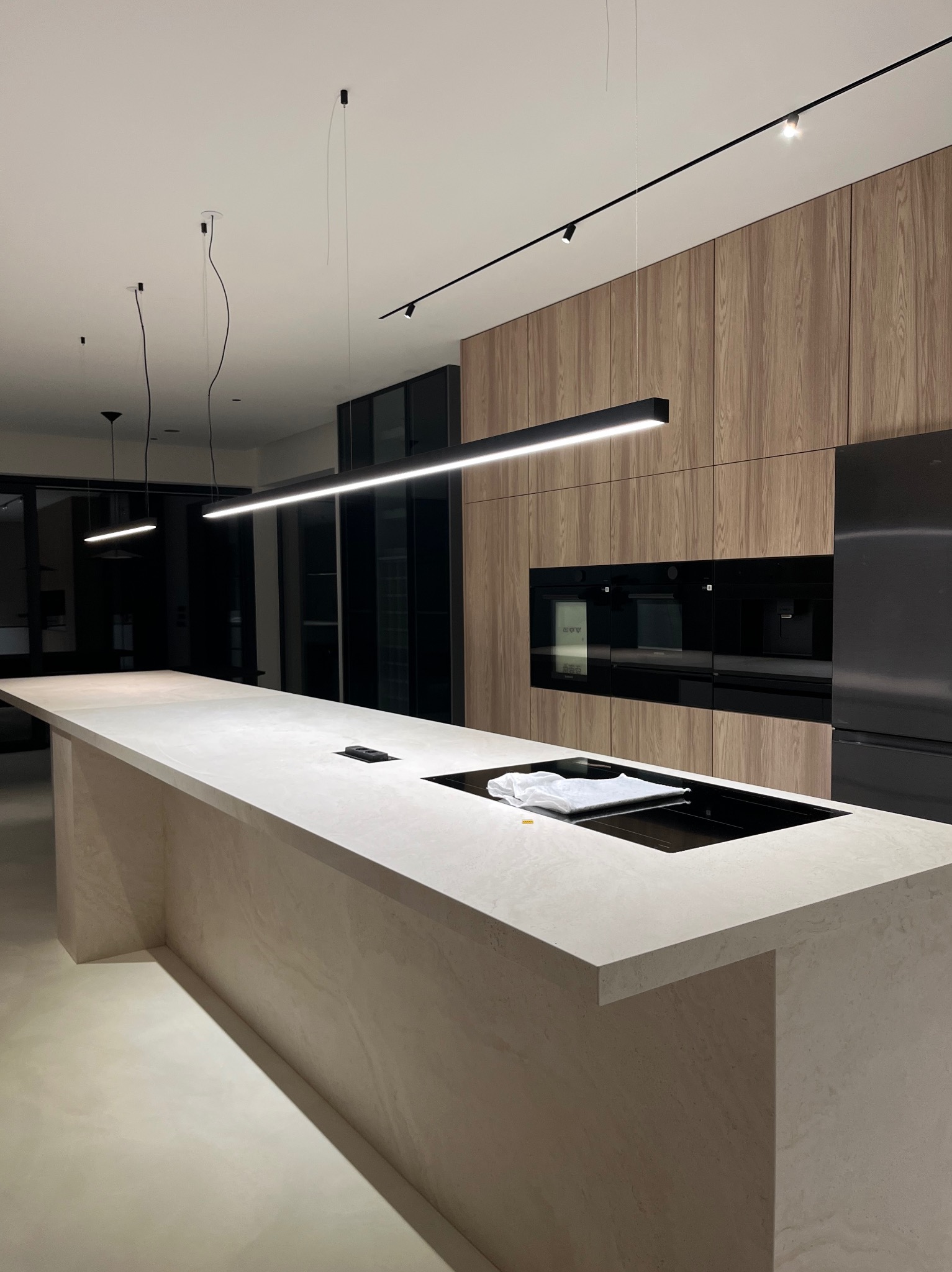 Nowoczesna kuchnia z wyspą i drewnianymi szafkami. Na blacie leży biała ściereczka. Oświetlenie punktowe i wiszące lampy. Minimalistyczny design.