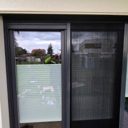 RollaTech Patryk Sadowski - Zamontowana moskitiera przesuwna w oknie tarasowym, widoczna siatka w czarnej ramie. W tle odbicie fotografa robiącego zdjęcie.