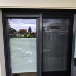 Zamontowana moskitiera przesuwna w oknie tarasowym, widoczna siatka w czarnej ramie. W tle odbicie fotografa robiącego zdjęcie.