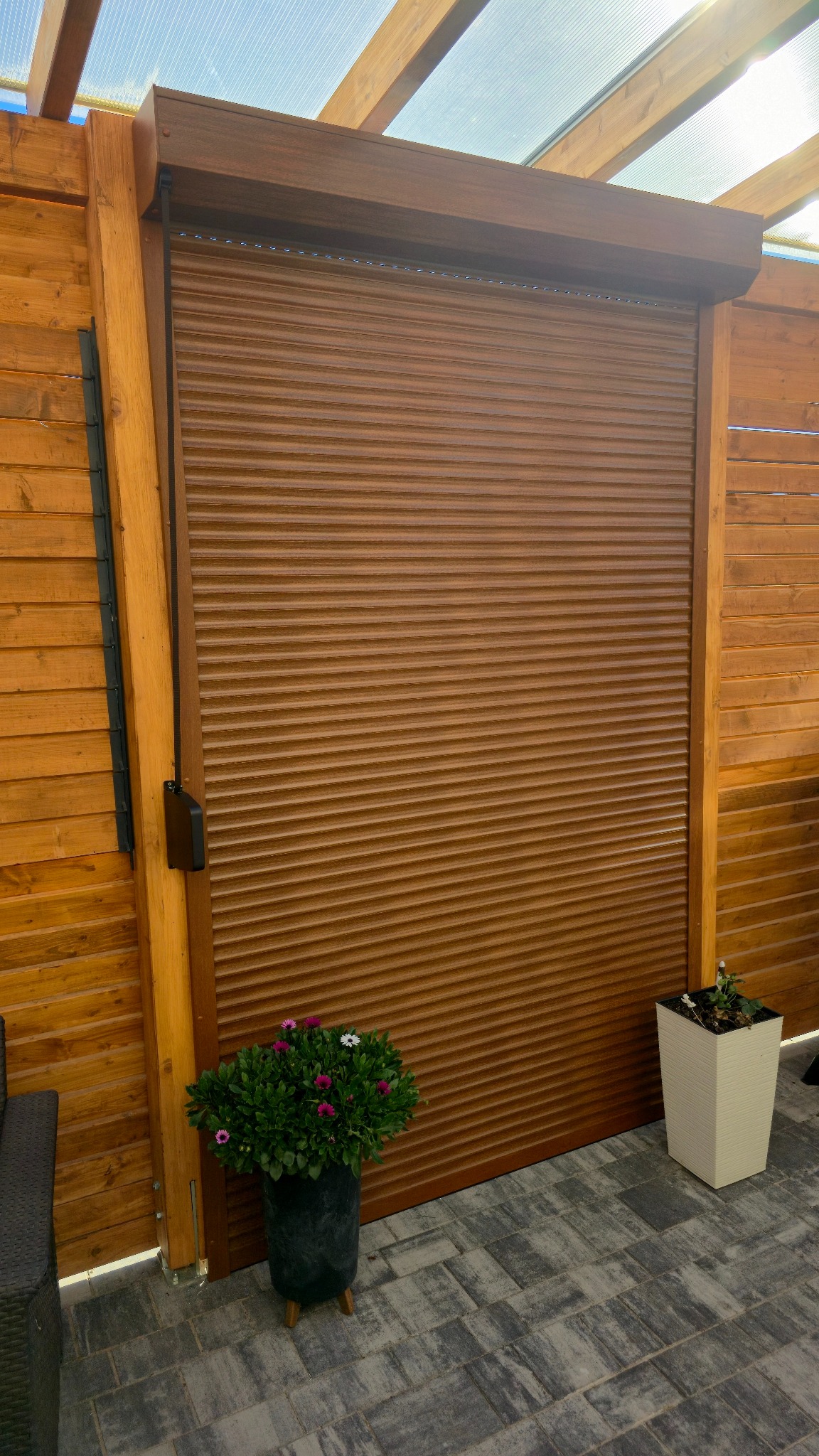 Zamknięta, brązowa roleta zewnętrzna na tle drewnianej konstrukcji z przezroczystym zadaszeniem. U dołu donice z kwiatami na szarej kostce brukowej.