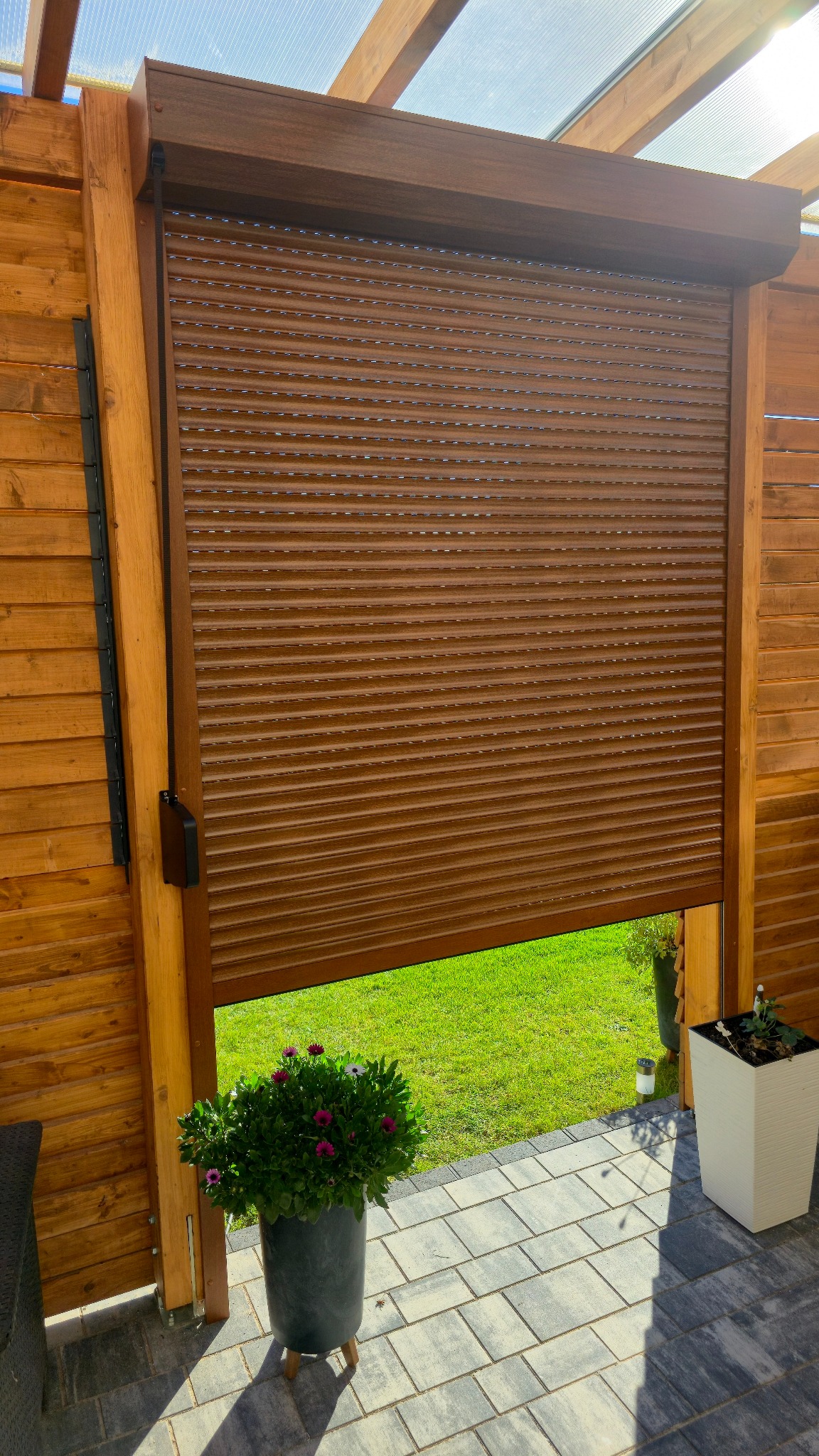 Częściowo opuszczona brązowa roleta zewnętrzna na drewnianej konstrukcji tarasu, z widokiem na trawnik i kwiaty w donicach. Słoneczny dzień.